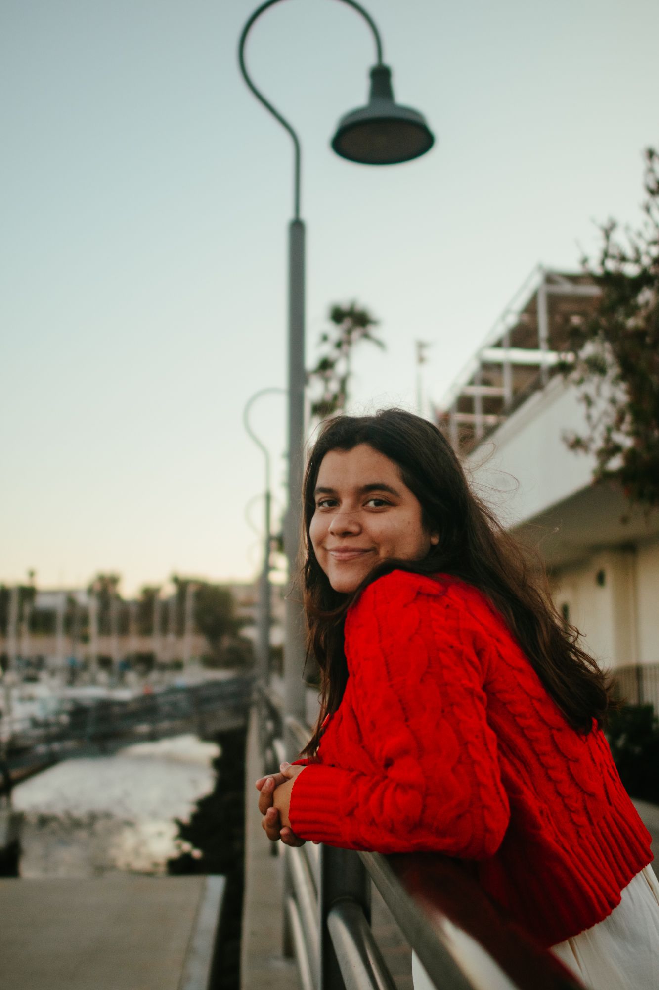 Young woman in red knit sweater leaning on railing at marina during golden hour sunset.