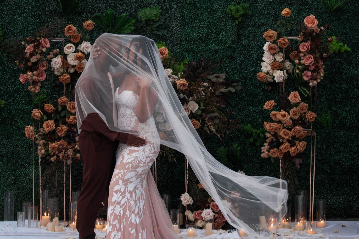 Bride and groom share romantic kiss under flowing veil at floral wedding arch with candlelight.
