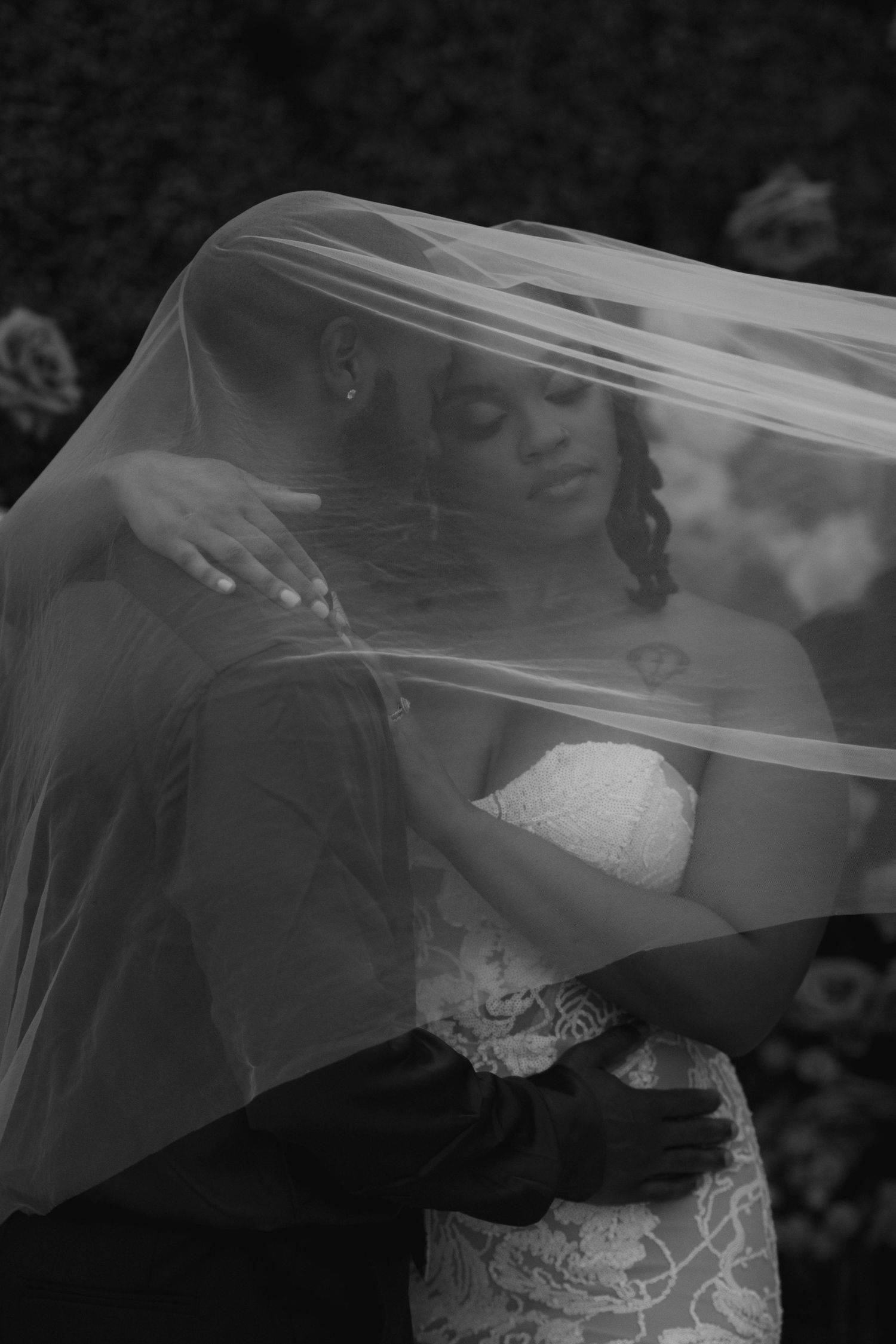Black and white photo of bride and groom embracing under a flowing veil outdoors.