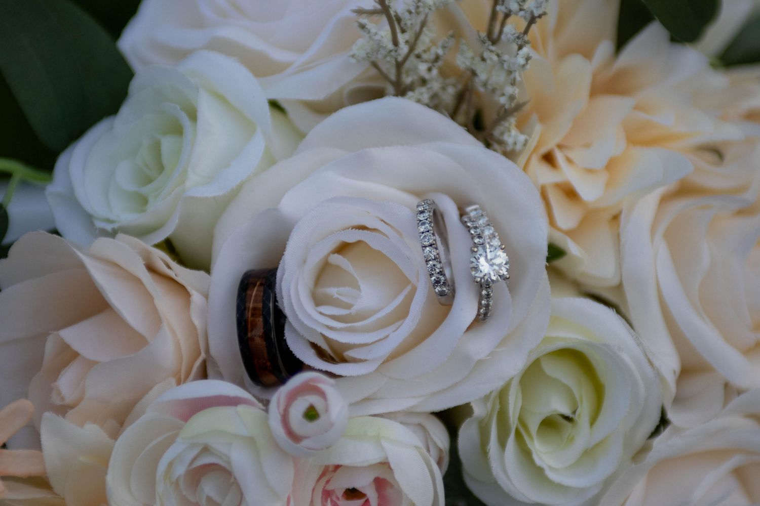 Diamond engagement ring and wedding bands nestled among white and blush roses in a bridal bouquet.