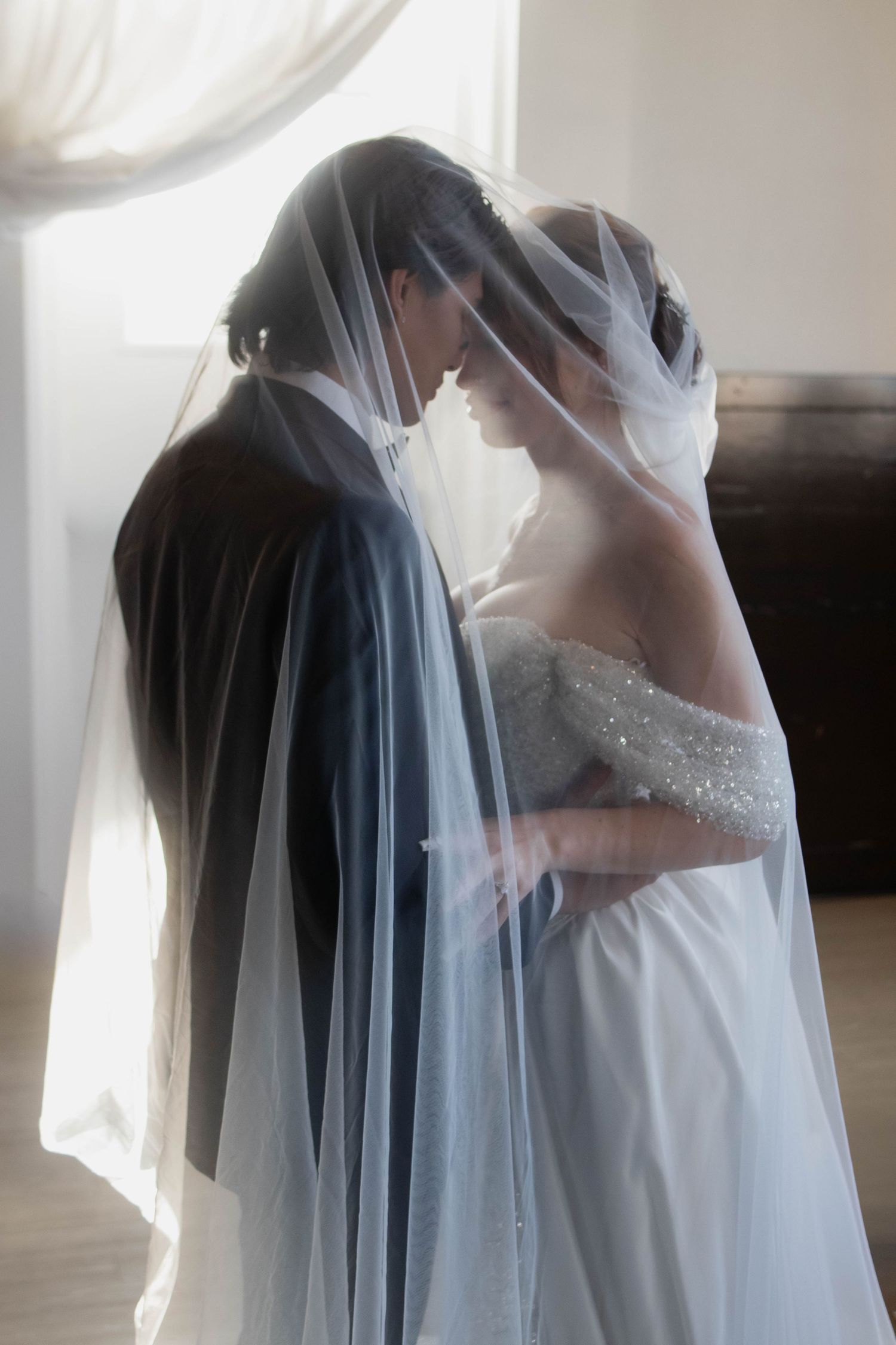Bride and groom sharing an intimate moment under a sheer wedding veil, soft backlit window glow.