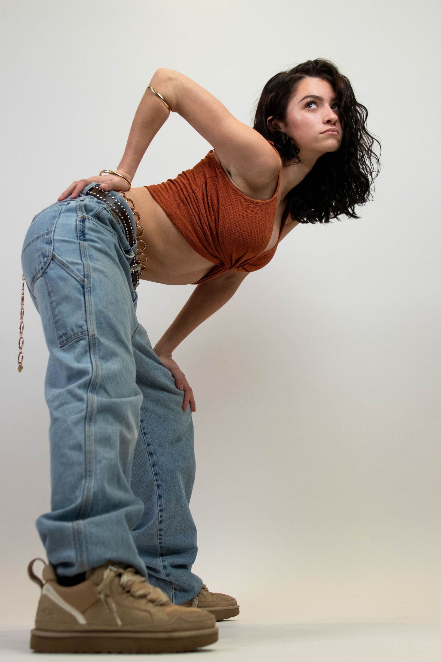 Young woman posing in baggy jeans, rust crop top, and sneakers on white background.