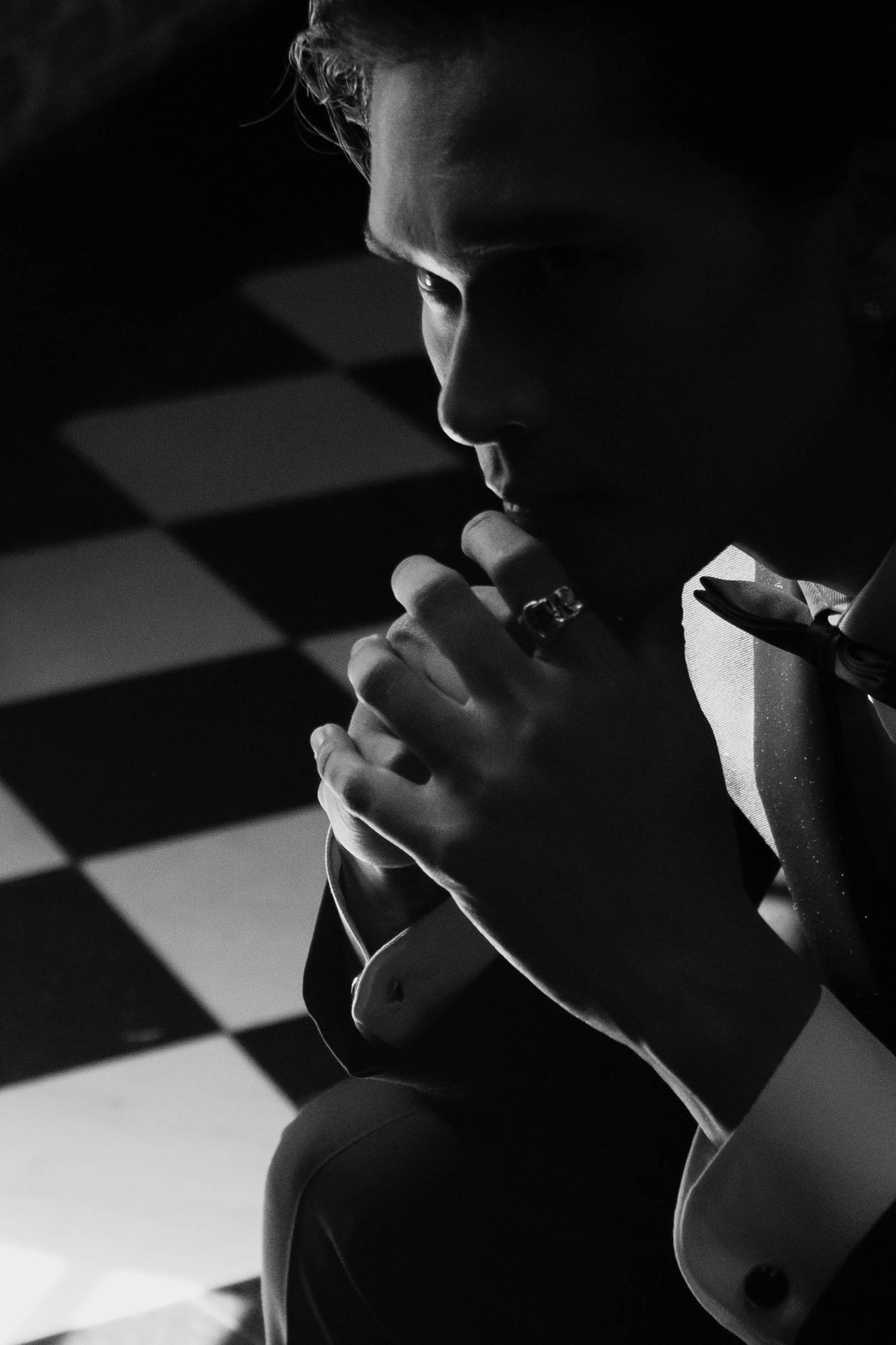 Black and white photo of a man in a suit with cufflinks, hands clasped thoughtfully near his lips on a checkered floor.