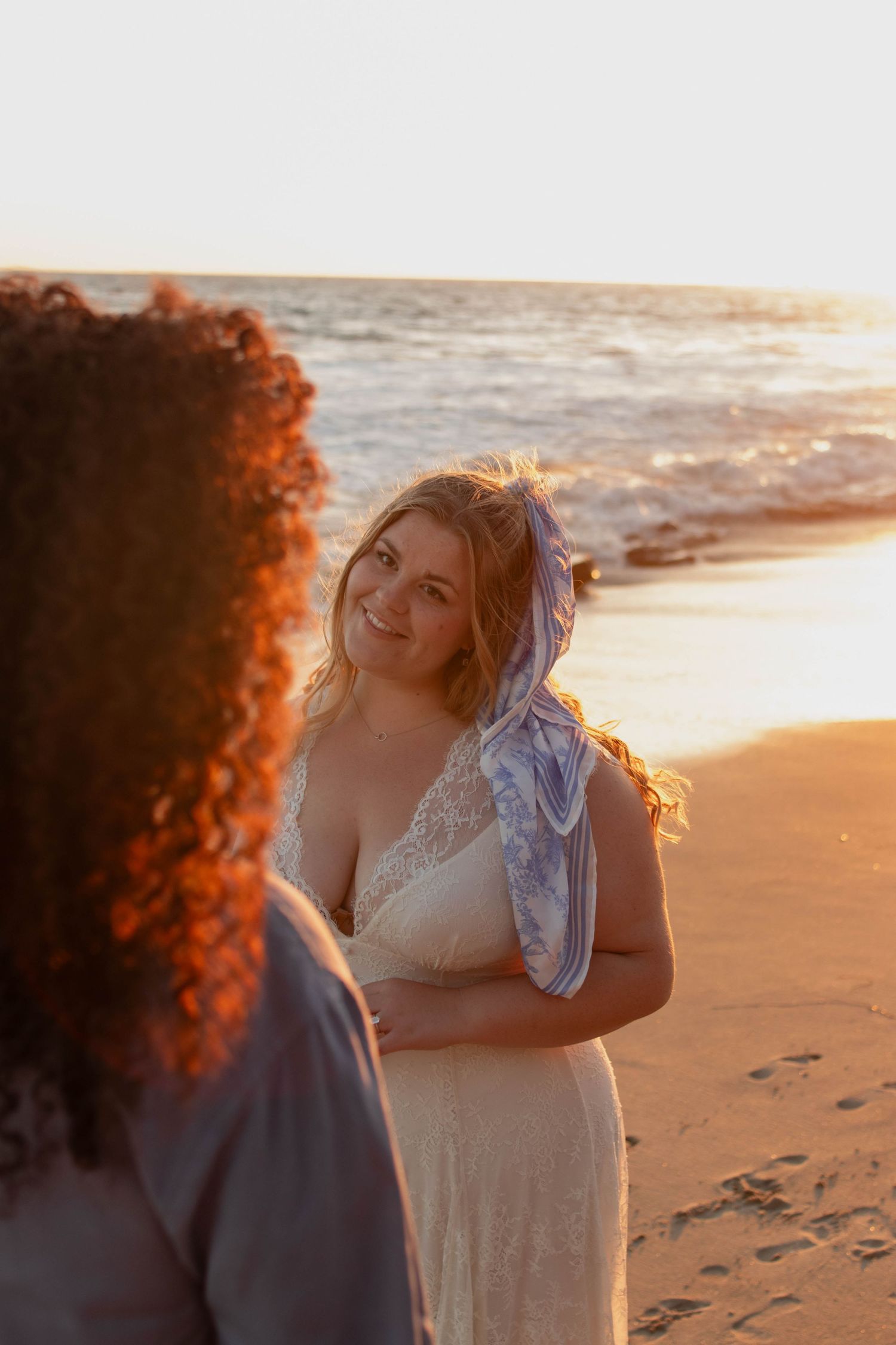 Two women share a romantic beach moment at golden sunset, one in a white lace dress with a blue scarf.