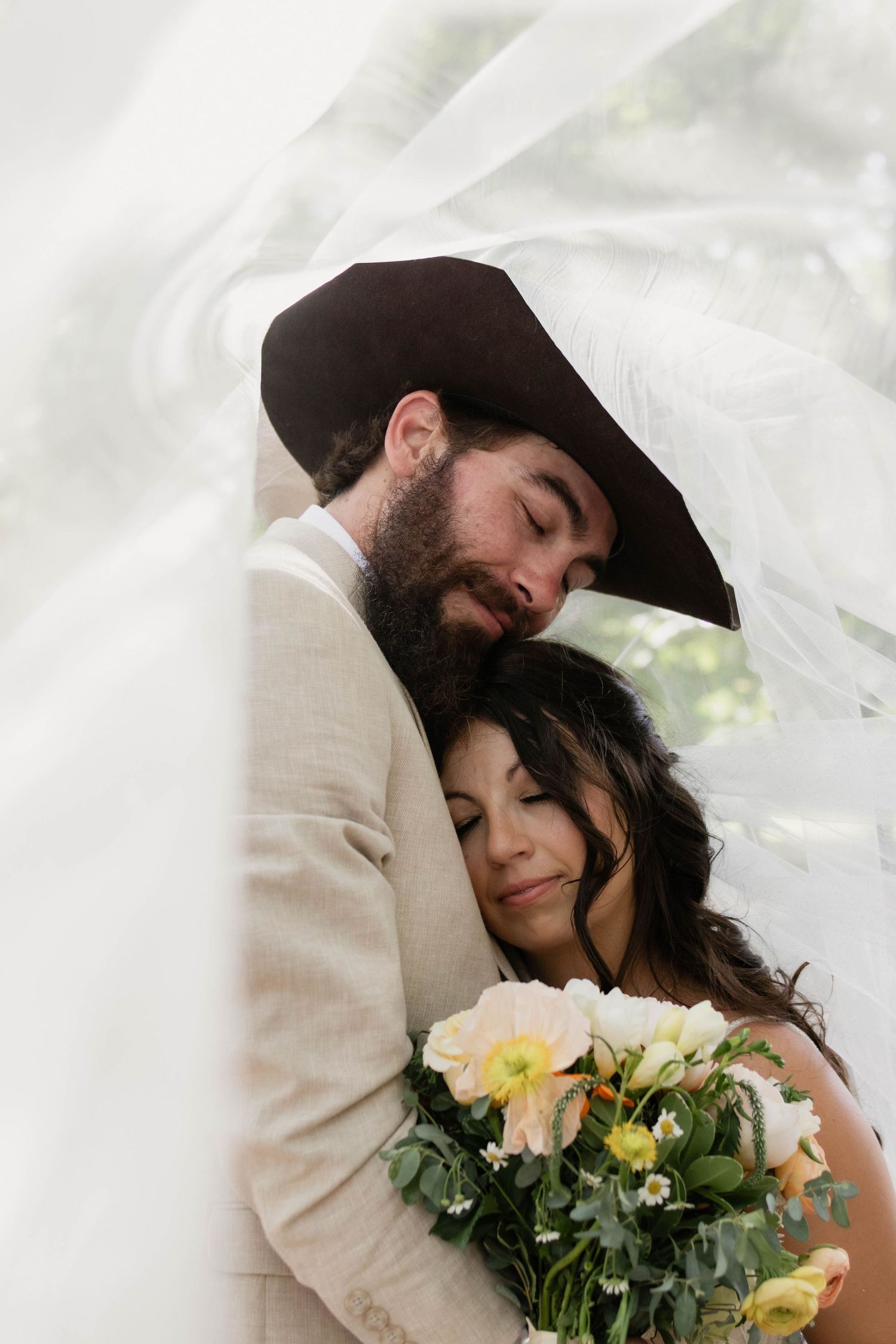 Groom in tan suit and cowboy hat embracing bride holding yellow and white bouquet under veil.