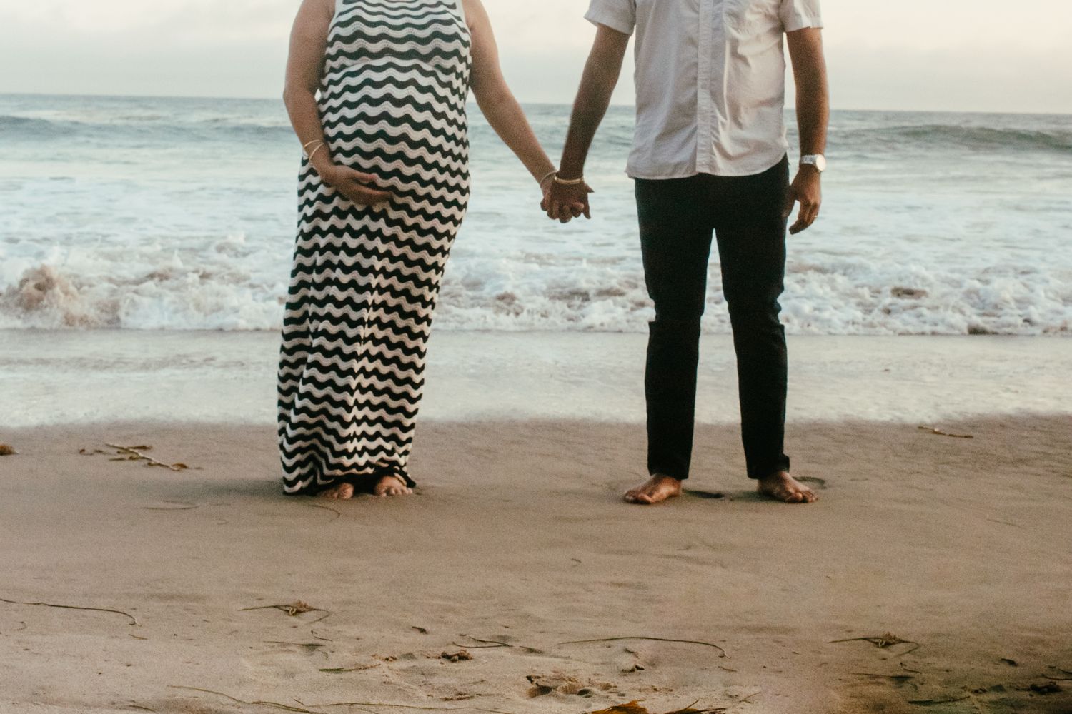 Pregnant woman in chevron maxi dress holding hands with partner while standing barefoot on sandy beach.