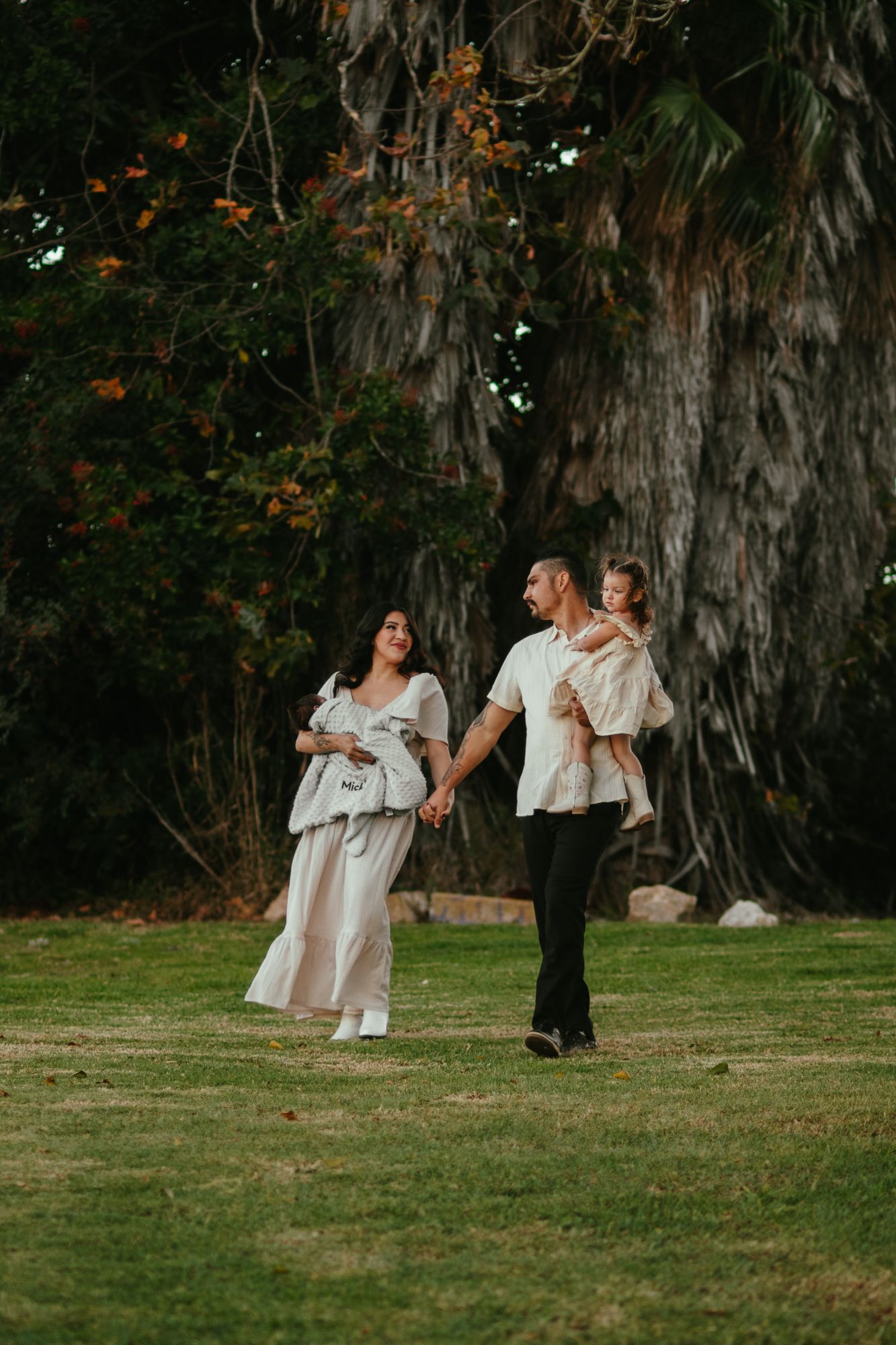 Happy family with baby walking hand in hand on green lawn in front of large tropical trees.