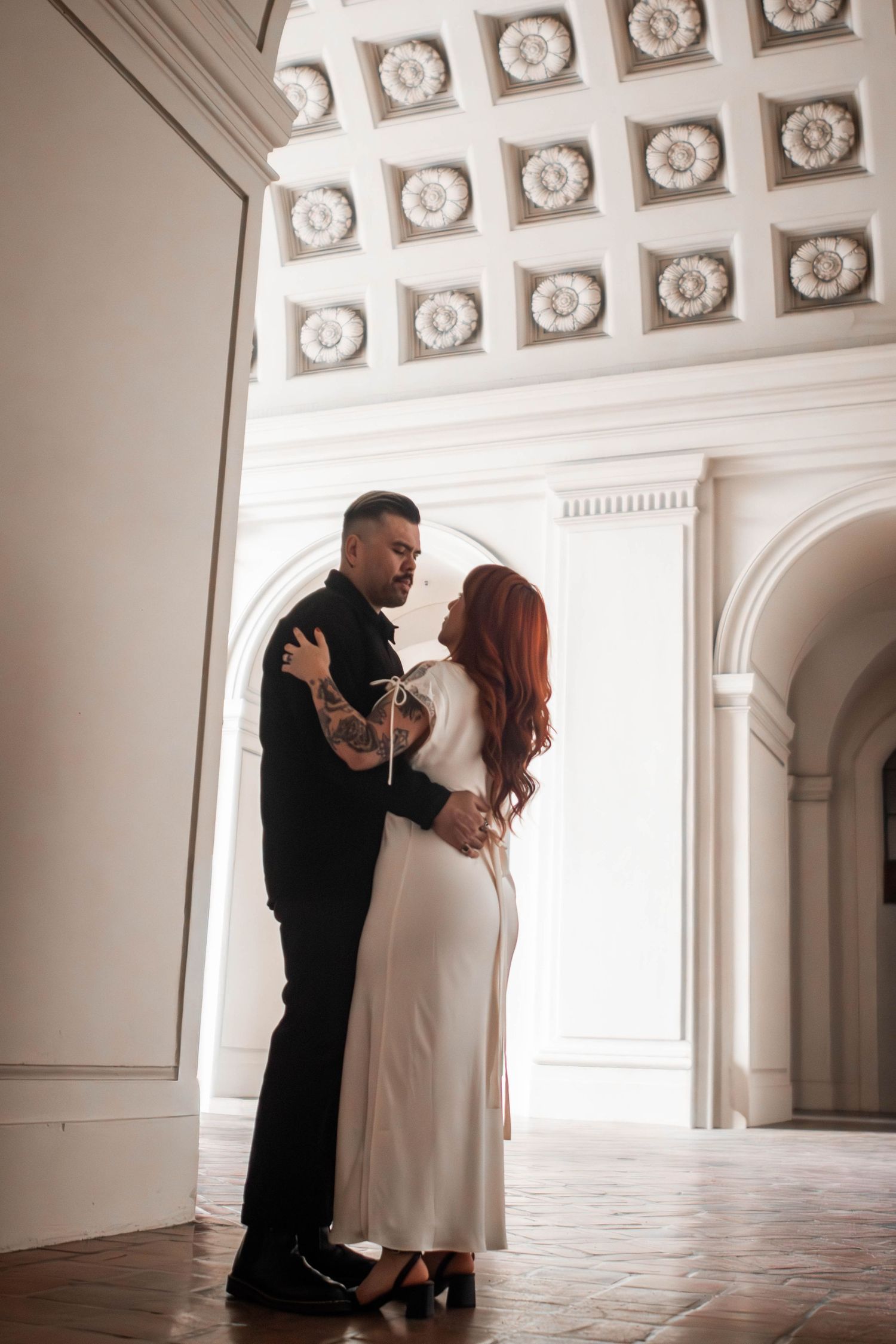 Couple sharing a romantic embrace inside an elegant white hall with ornate coffered ceiling.