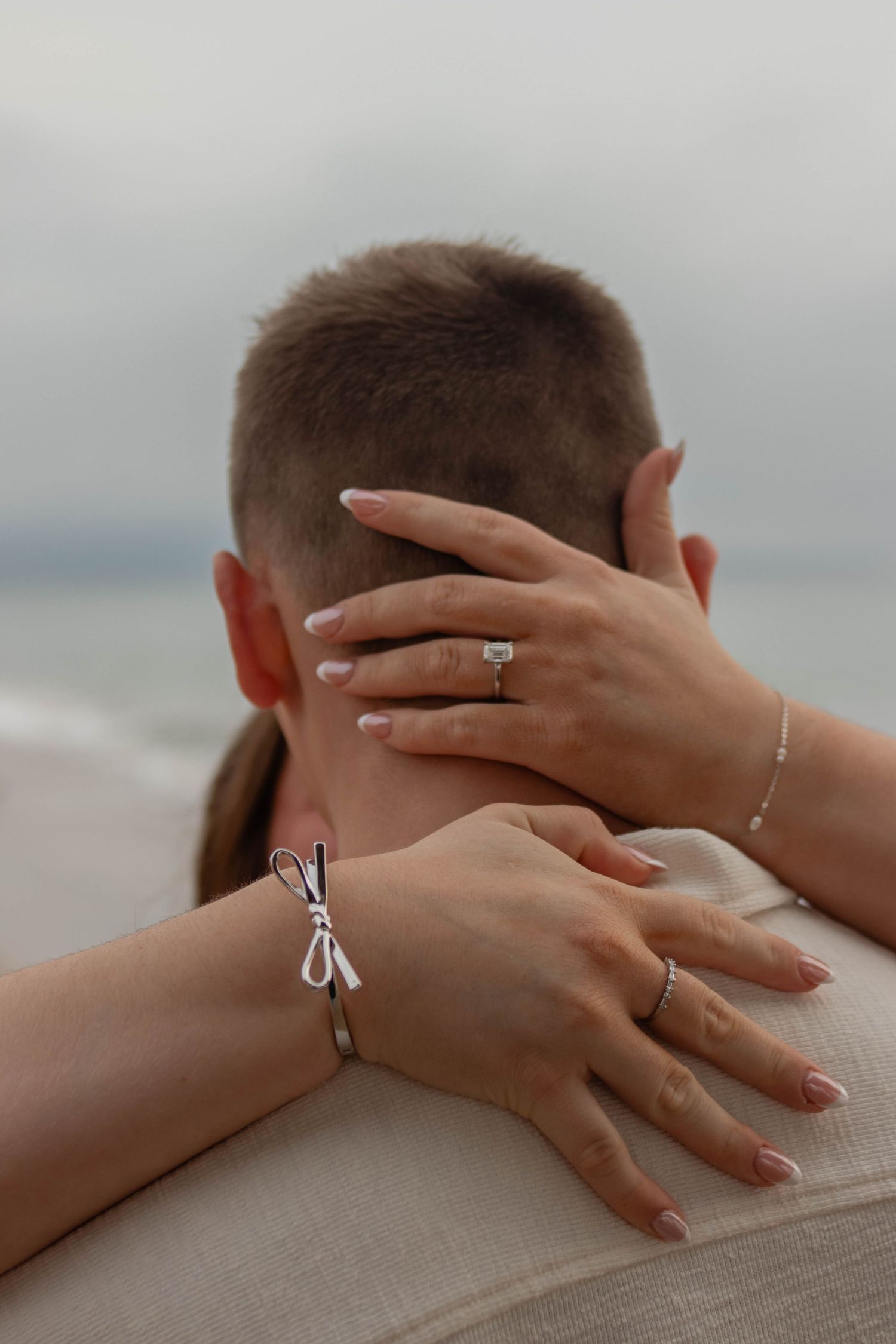 Close-up of womans hands with diamond engagement ring embracing partner on cloudy beach setting.