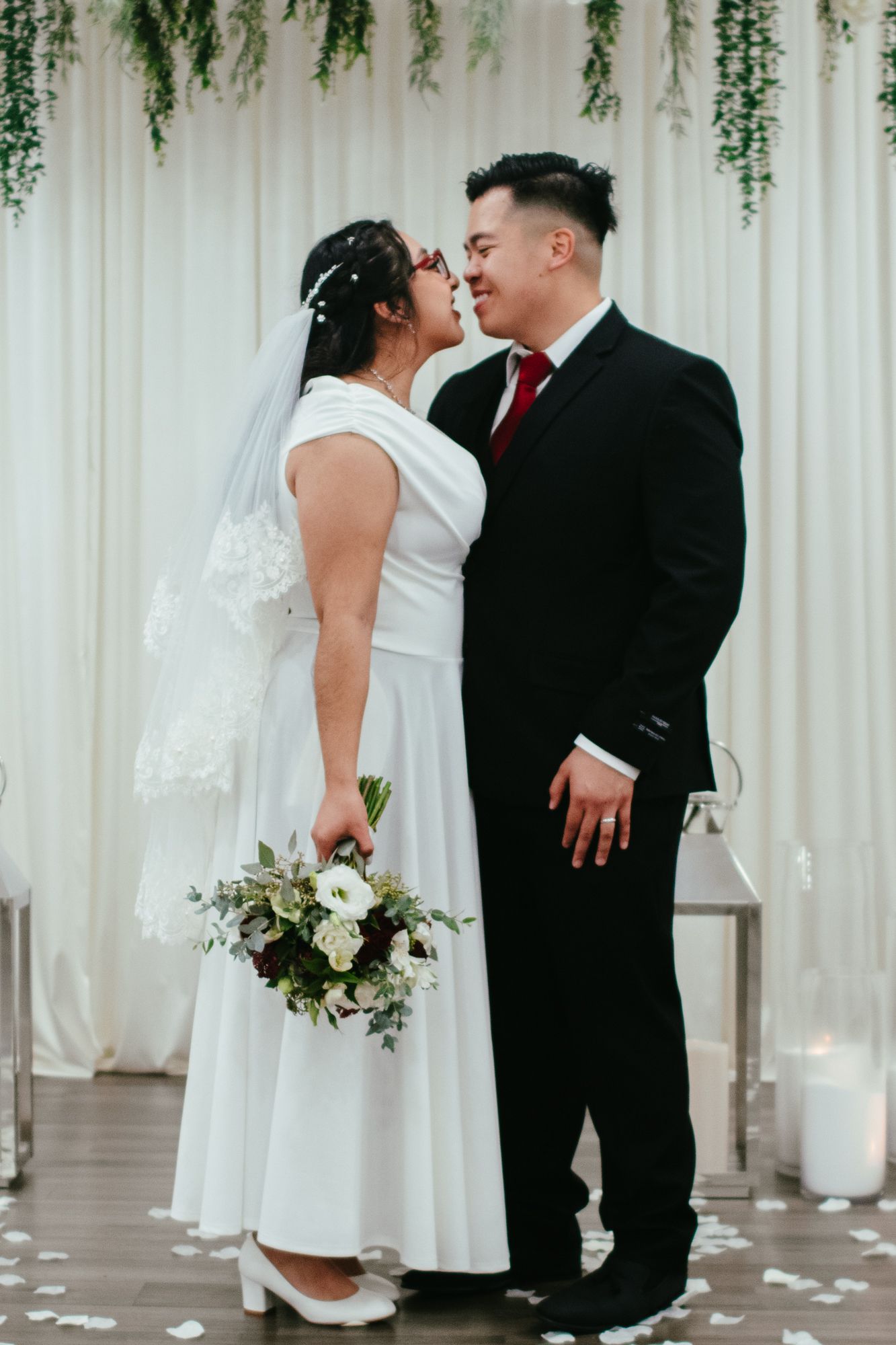 Asian bride and groom share first kiss at altar with white draping and greenery backdrop.