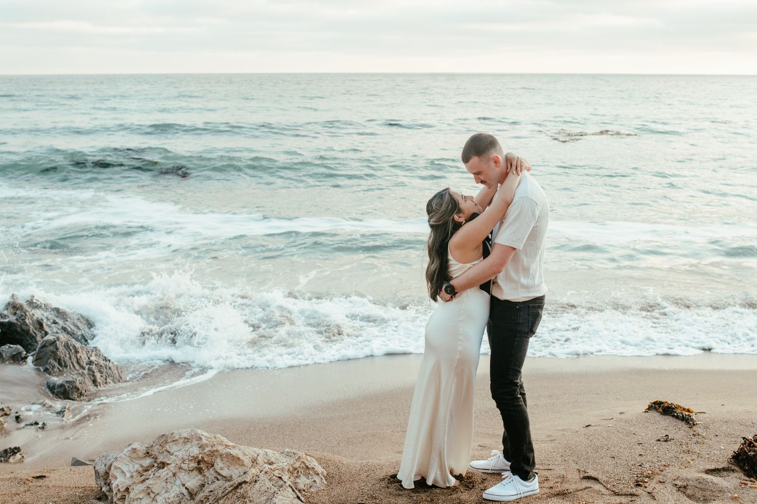 Romantic couple embracing on a rocky beach with ocean waves crashing behind them at sunset.