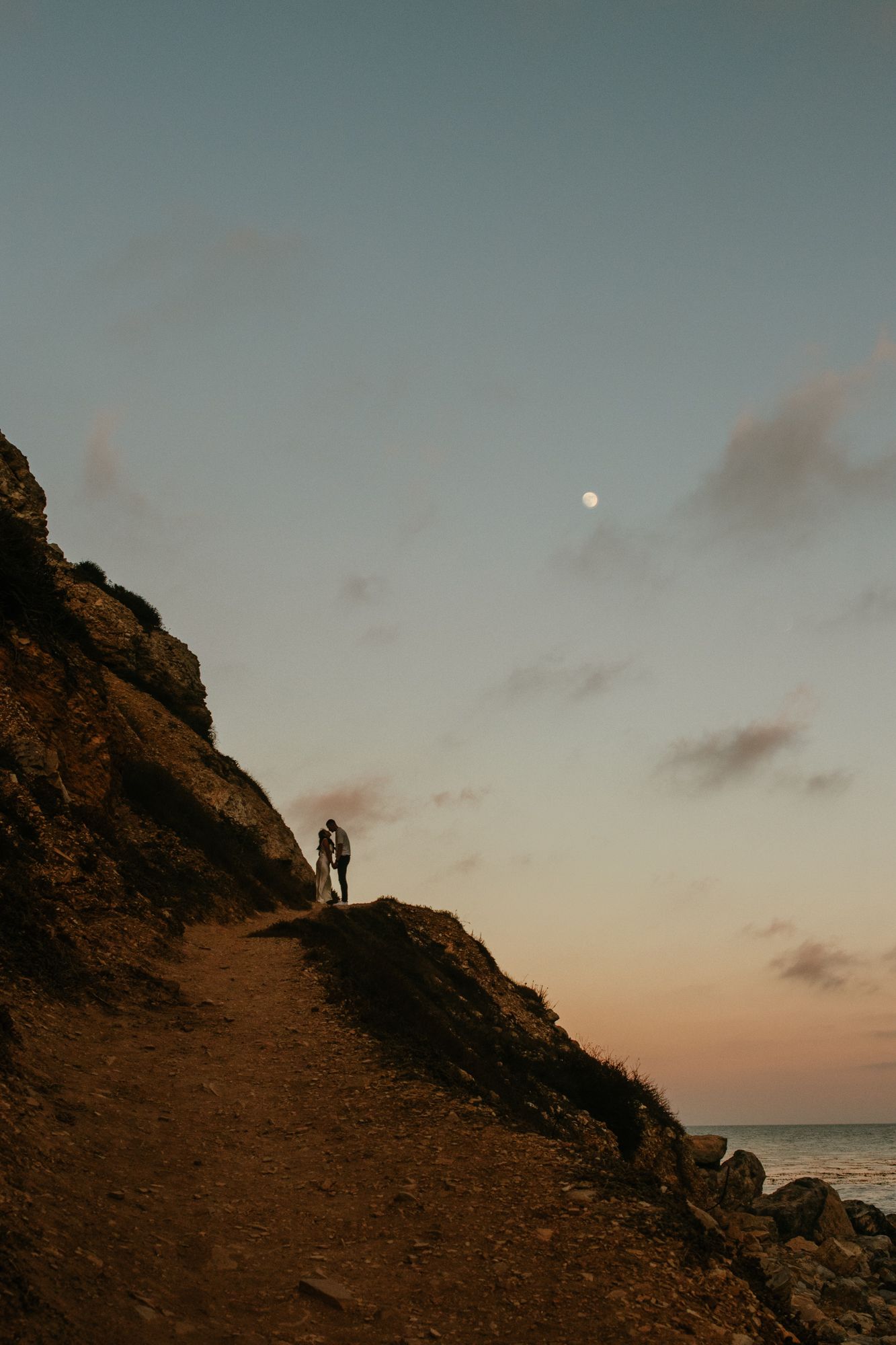 Couple silhouetted on a rocky coastal cliff path at twilight with the moon visible in the pastel sky.