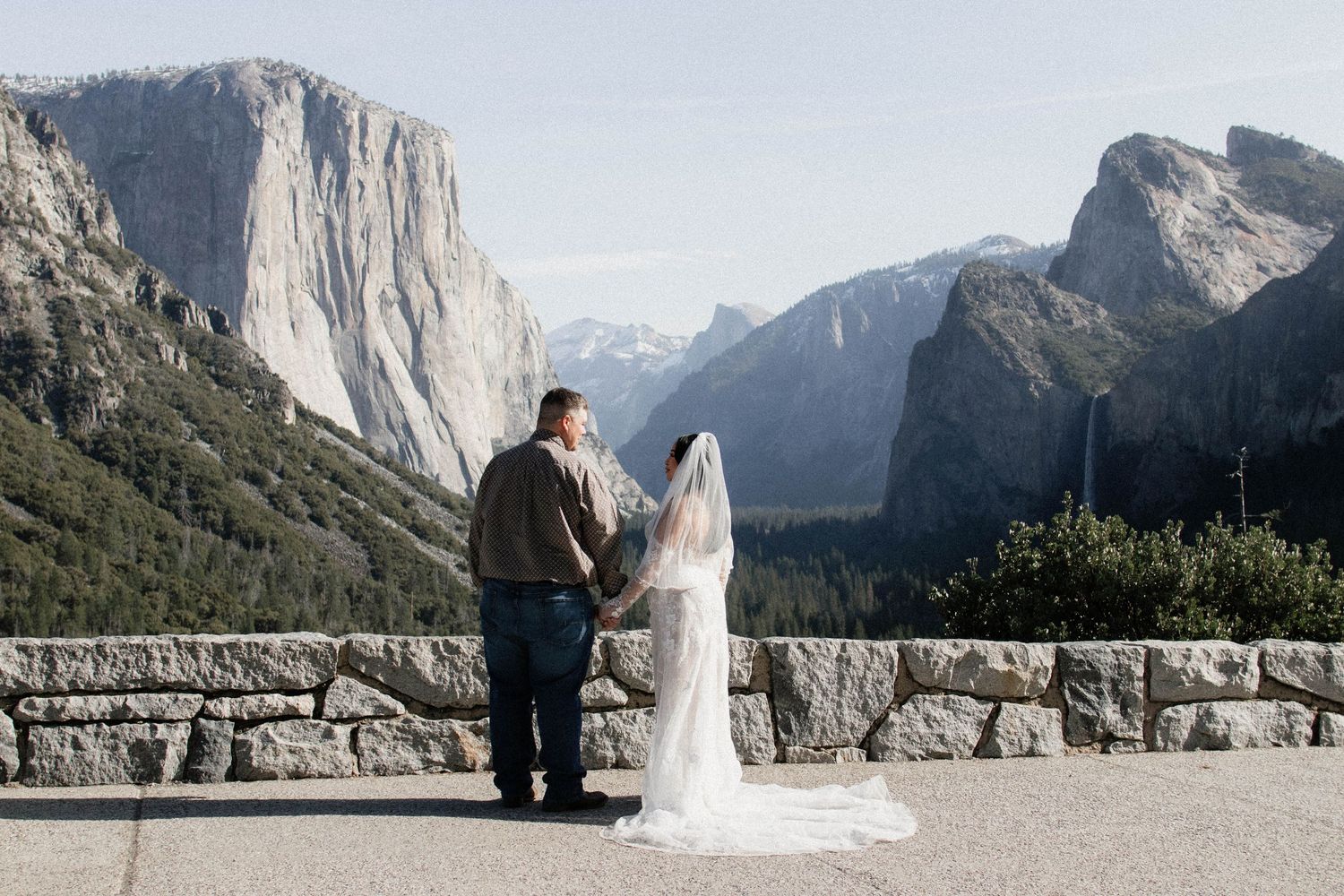 Bride and groom holding hands at Yosemite Valley overlook with stunning mountain backdrop.