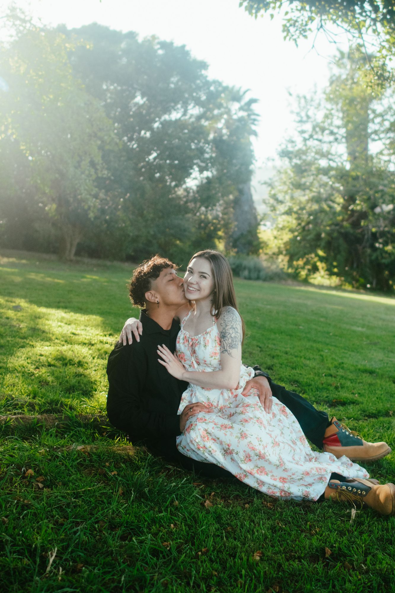 Happy couple sitting on green grass in sunlit park, man kissing woman's cheek during engagement shoot.