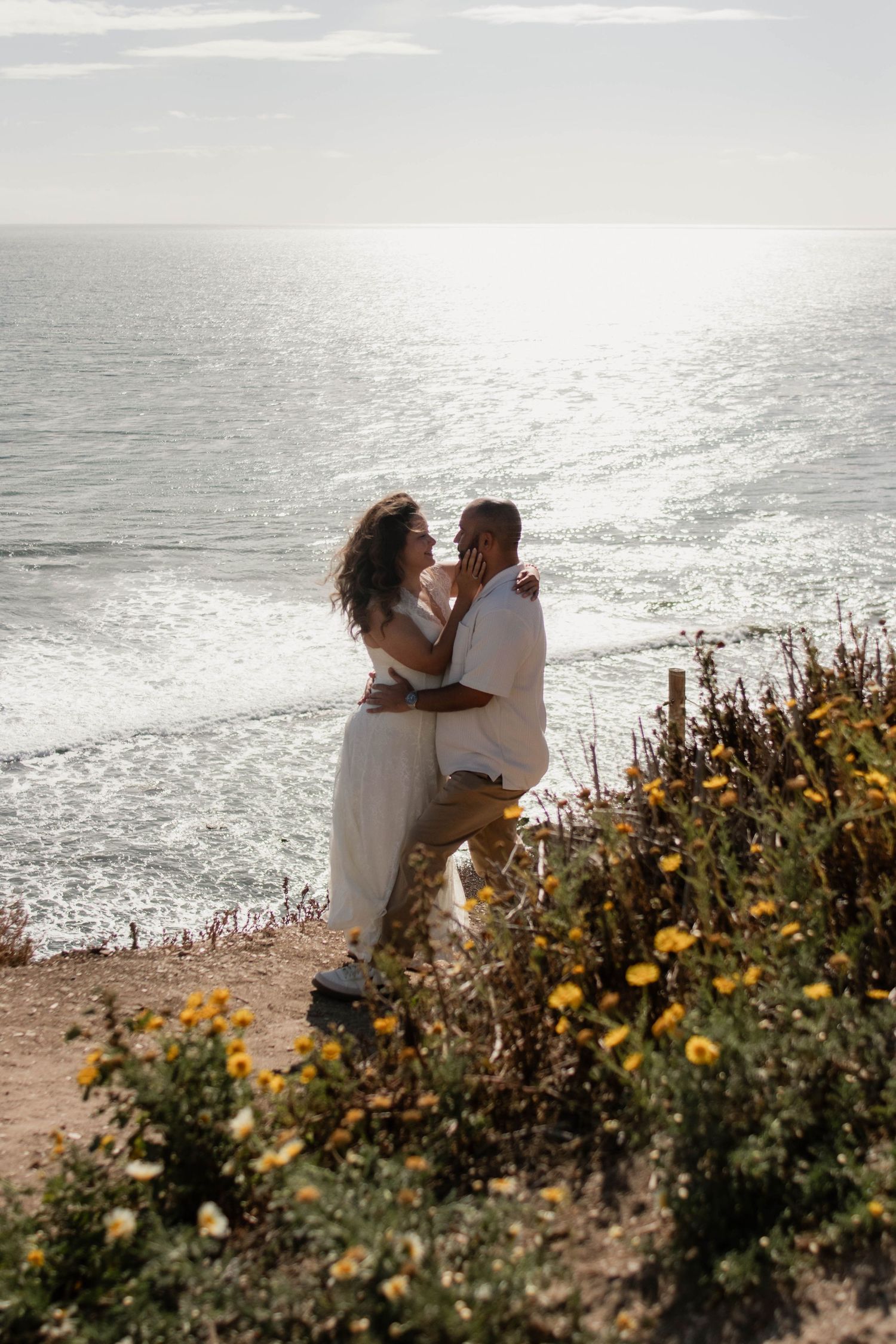 Couple embracing and kissing on a coastal cliffside with yellow wildflowers and sparkling ocean views.