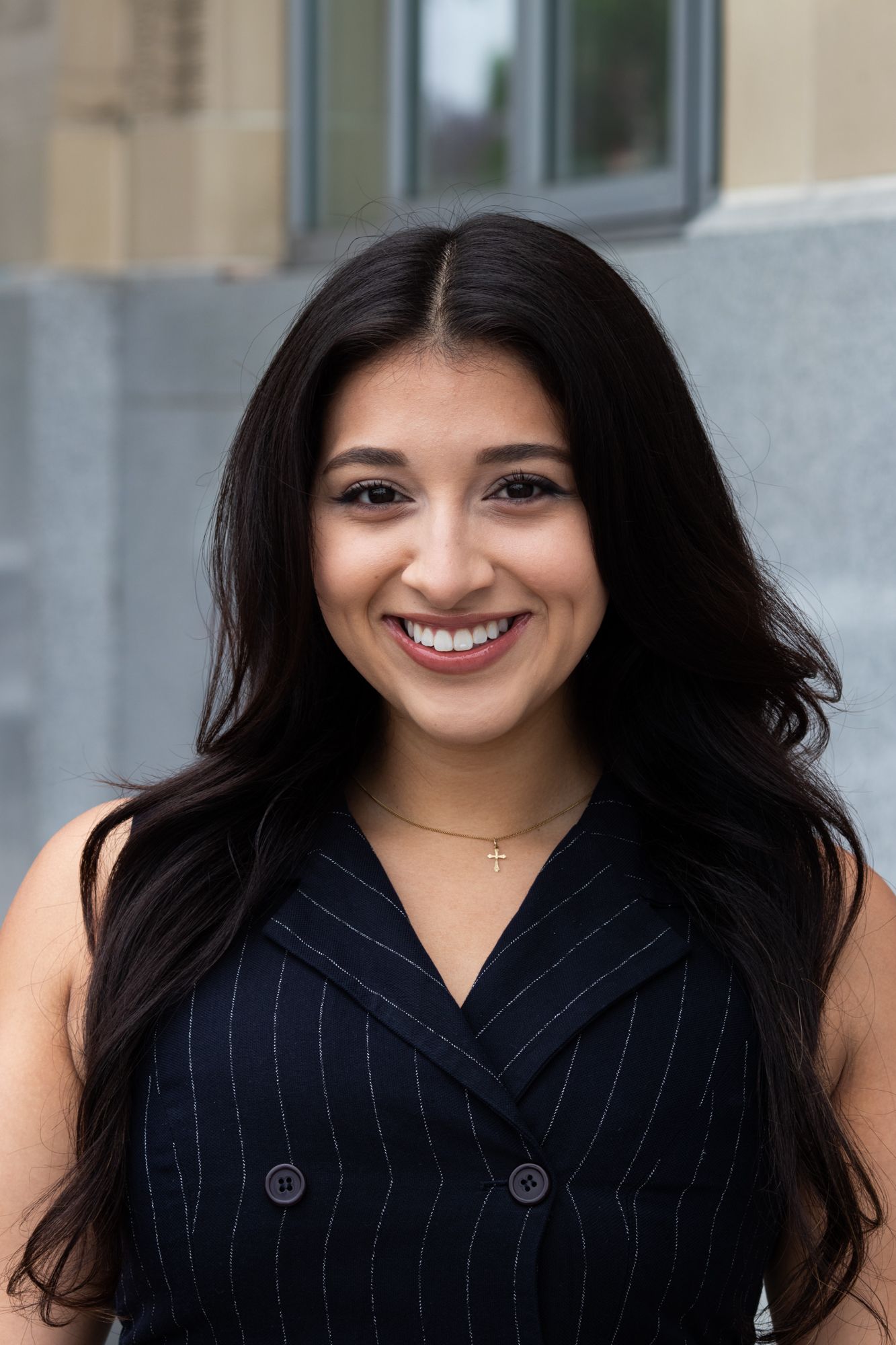 Smiling young woman with long dark hair wearing a navy pinstripe vest outdoors.