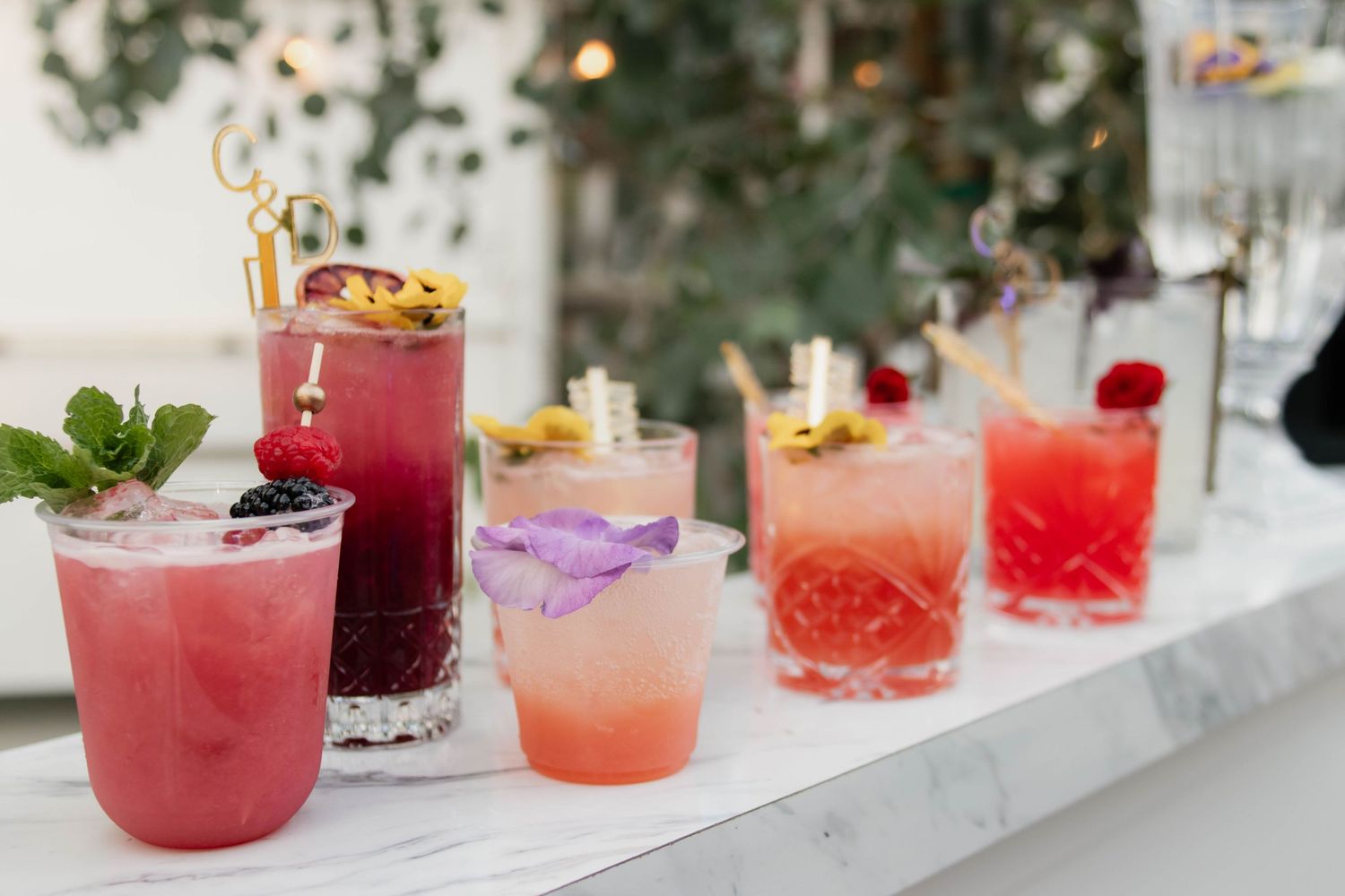 Colorful signature cocktails with floral garnishes displayed on white marble bar at wedding reception.