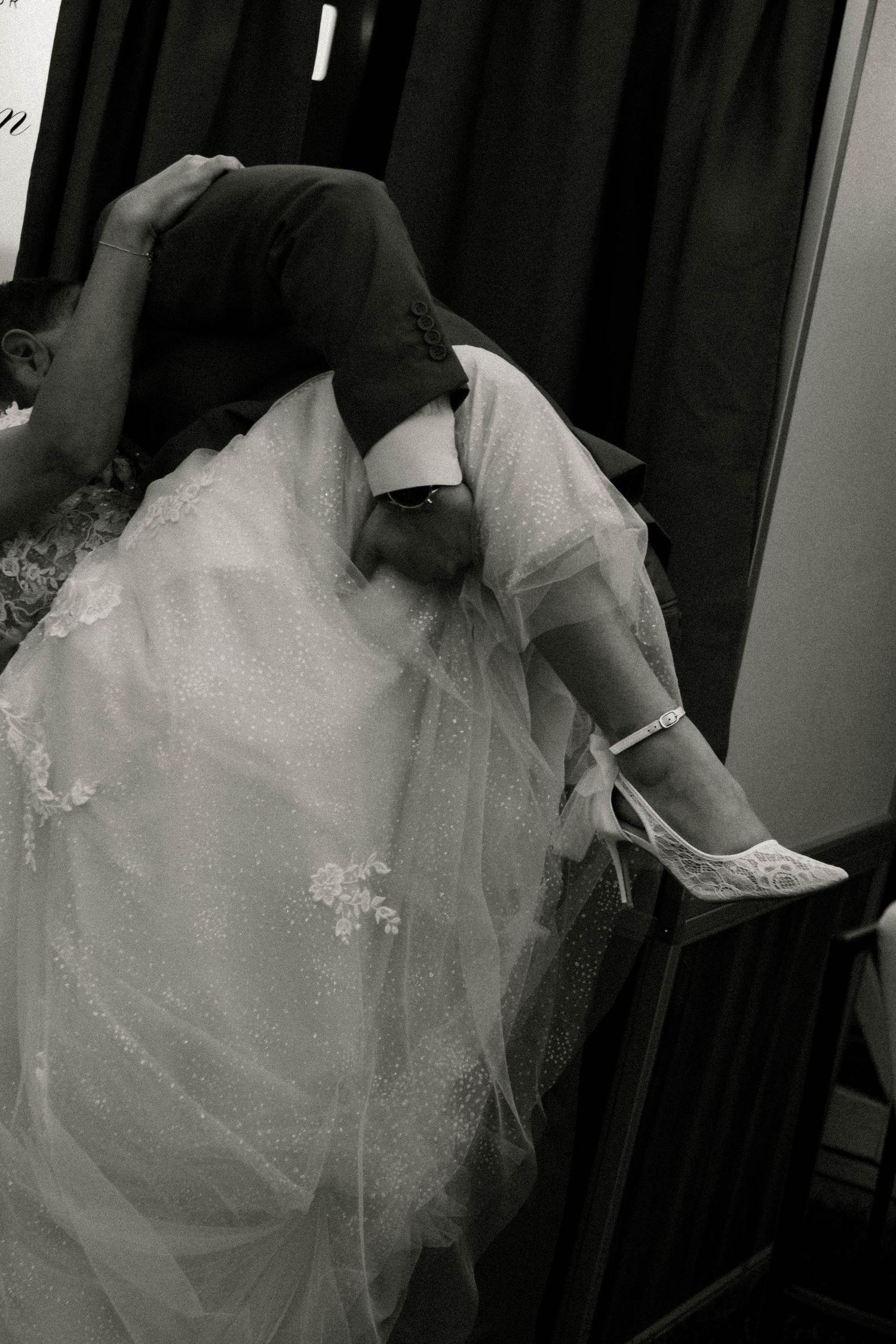 Black and white photo of a groom adjusting the brides white lace wedding heels beneath her flowing gown.
