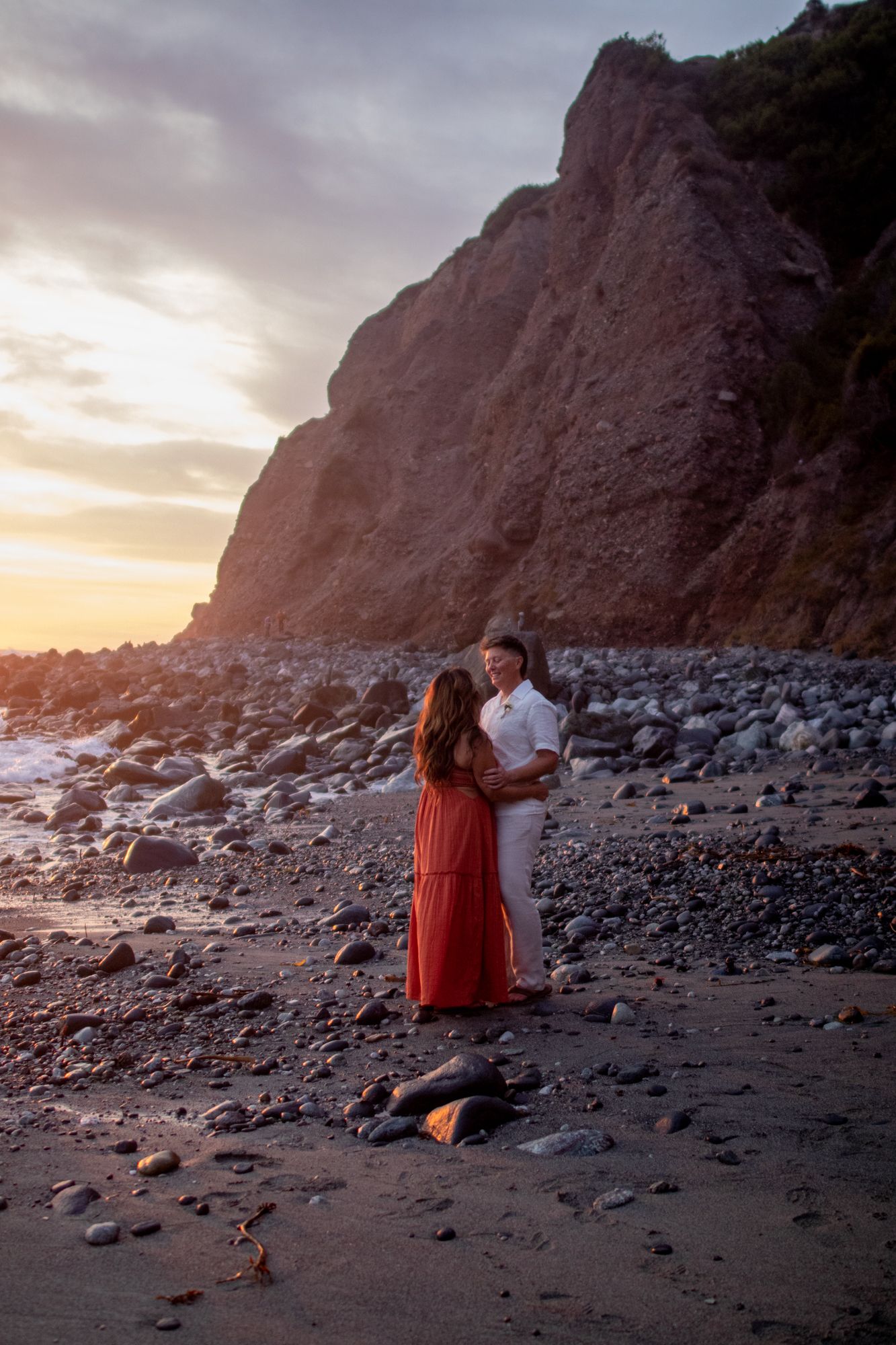 Couple embracing on a rocky beach at sunset, woman in red maxi dress and man in white shirt against dramatic cliff backdrop.