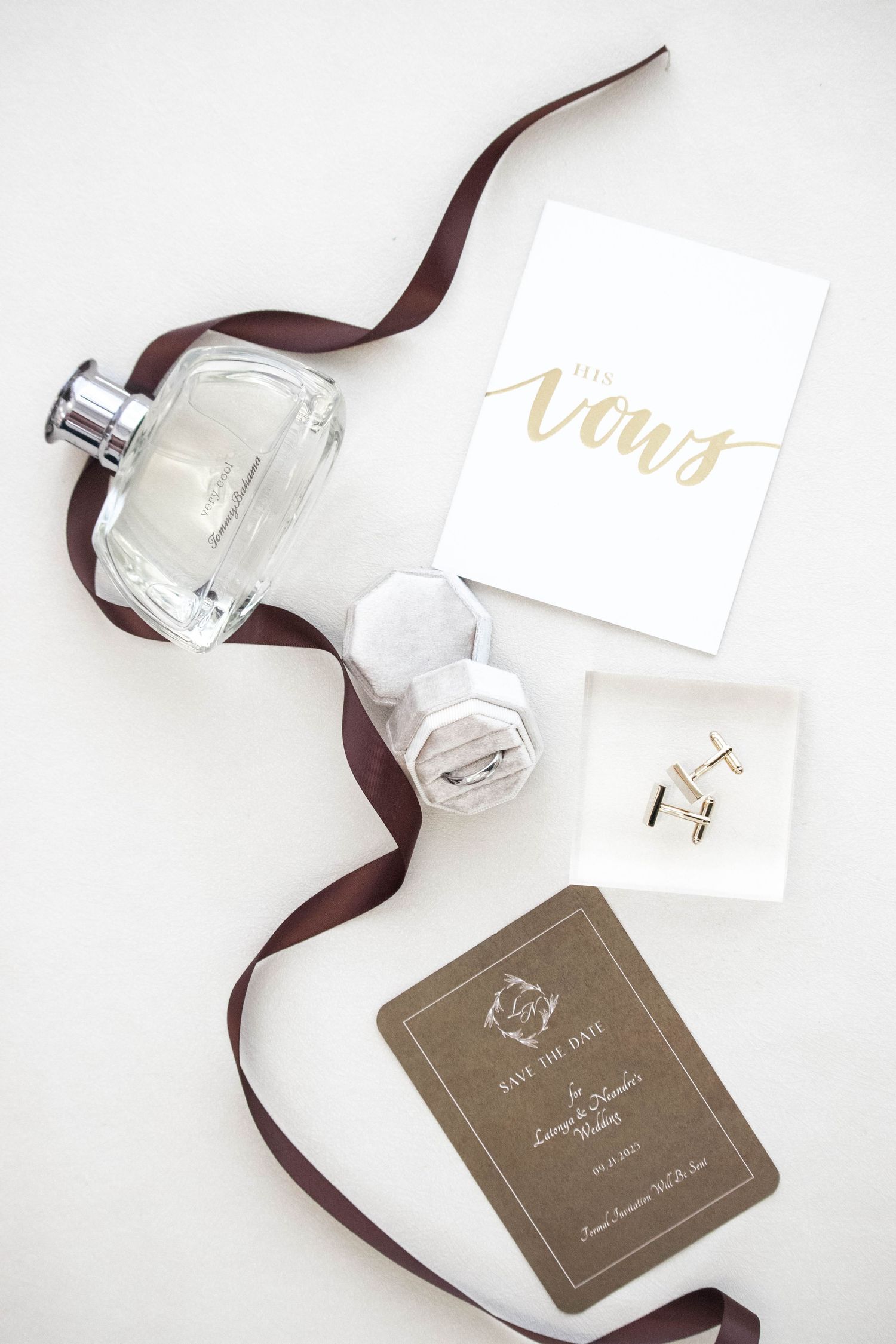 Wedding flat lay featuring perfume bottle, vow booklet, ring box, cufflinks, and save the date card with burgundy ribbon.