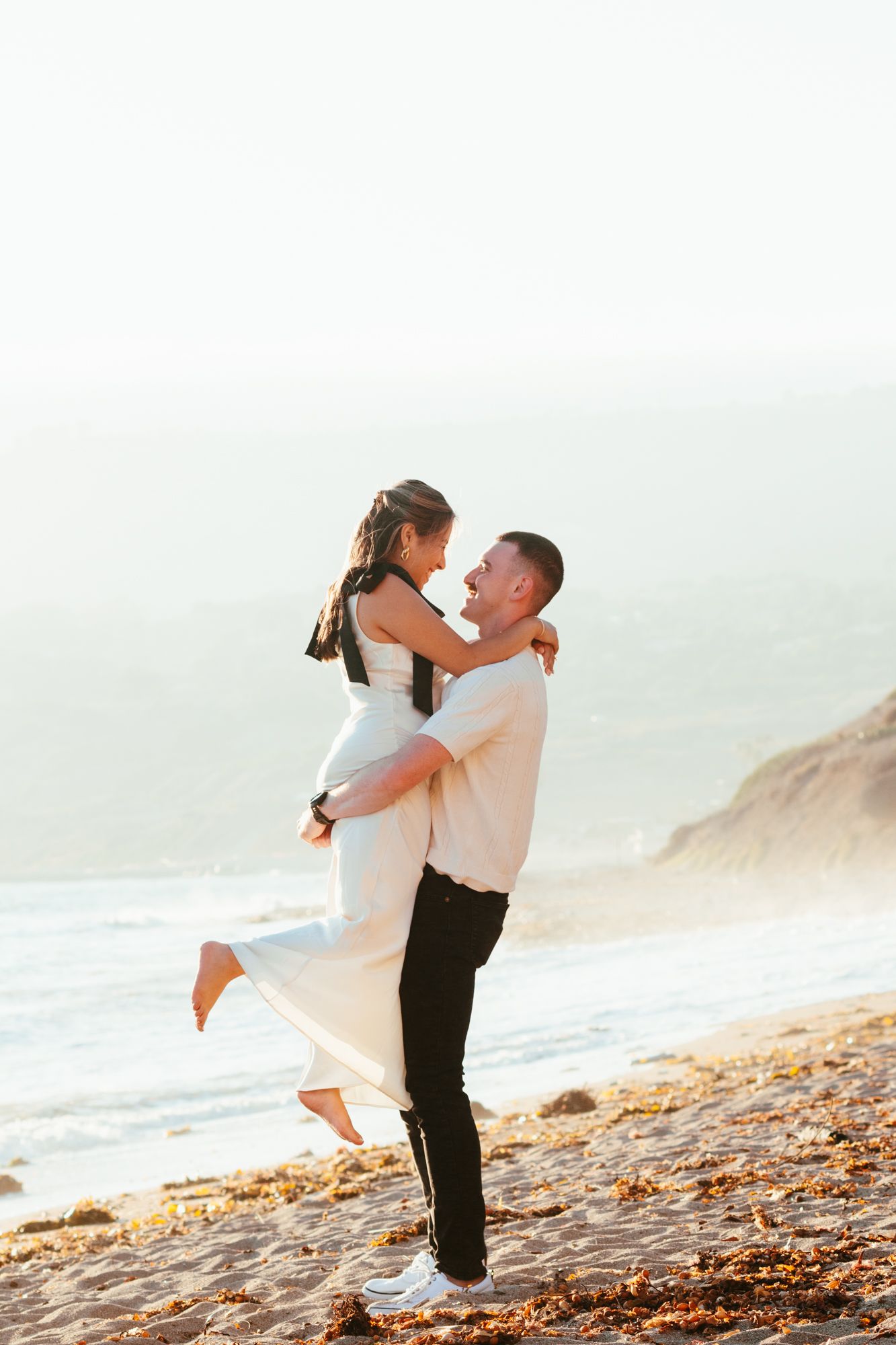 Groom lifting bride on sunny beach with ocean waves and cliffs in background.