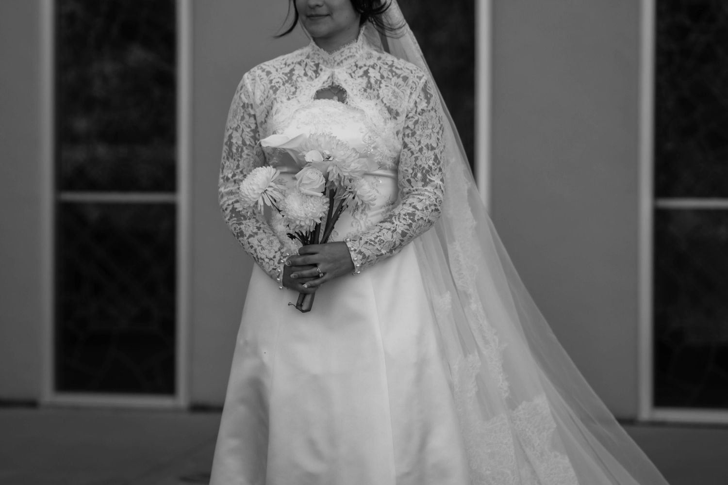 Bride in long-sleeve lace wedding gown with veil holding white bouquet, black and white photo.