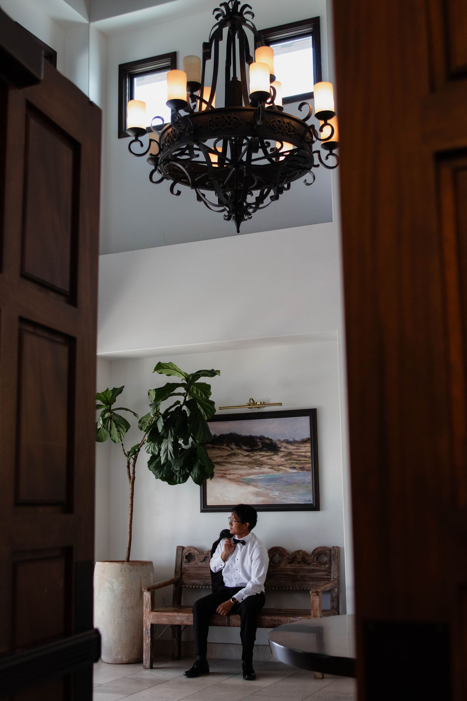 Man in white shirt sitting in elegant lobby with wrought iron chandelier, tropical plant, and landscape painting on wall.