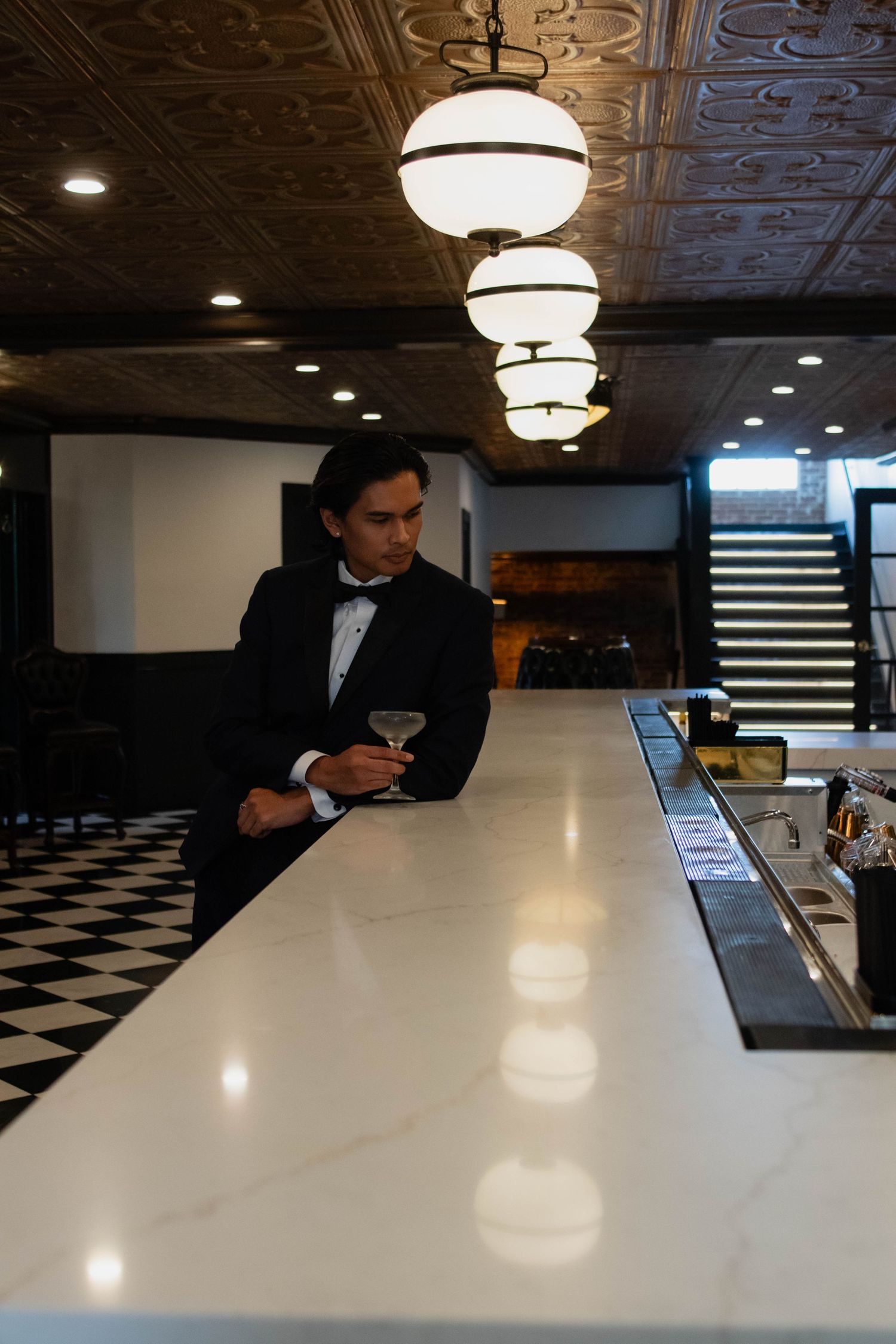 Elegant man in tuxedo holding martini glass at upscale bar with marble counter and decorative ceiling.