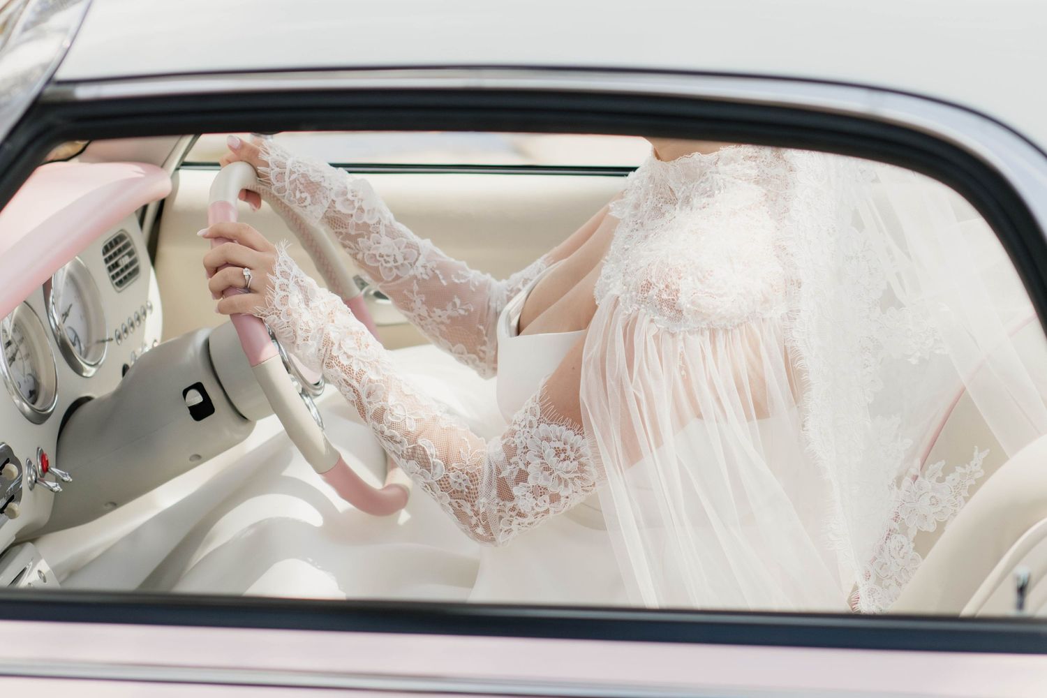 Bride in lace wedding dress and veil sitting in a white luxury car, holding the steering wheel.