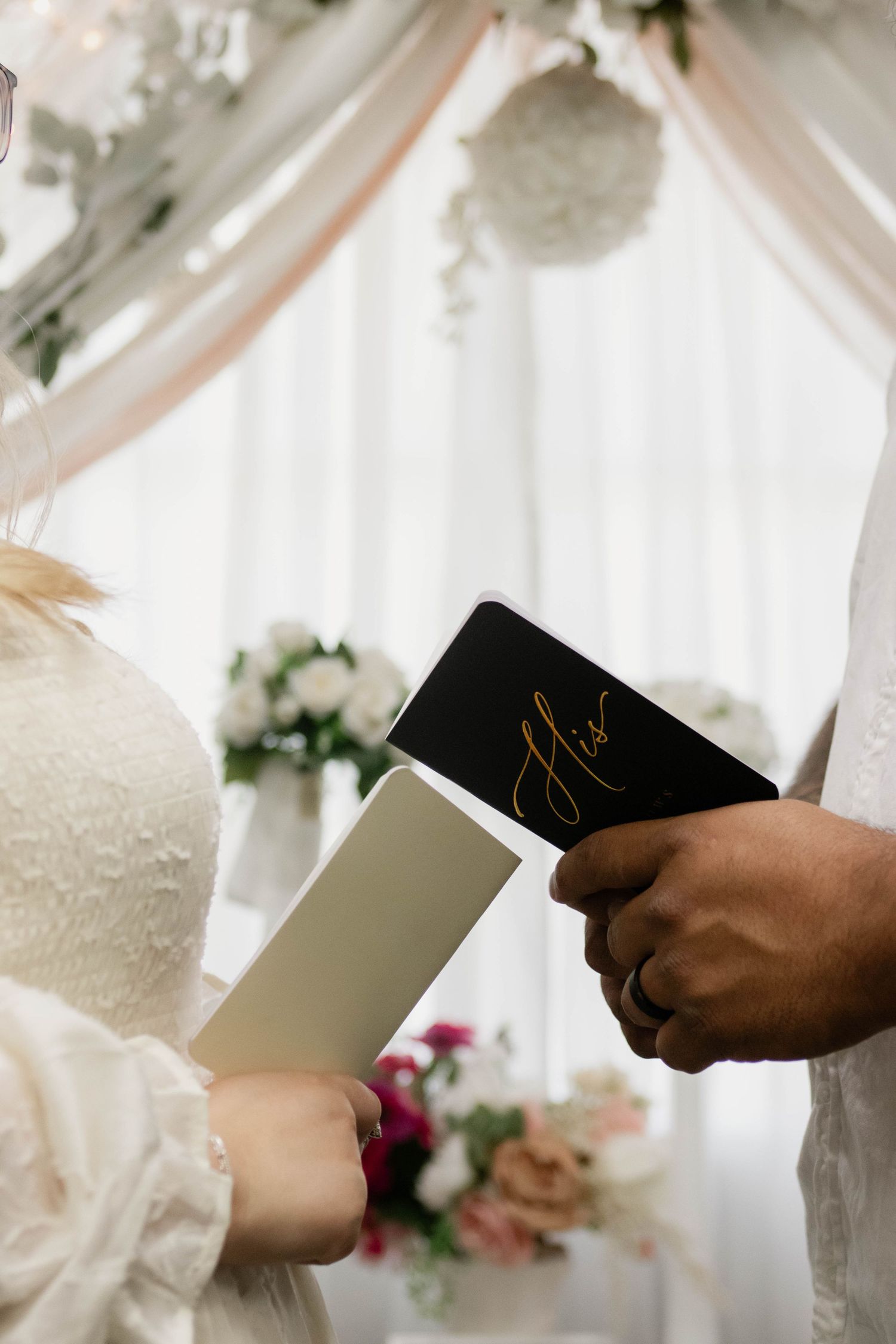 Couple exchanging wedding vows holding booklets at decorated altar with white draping and flowers.