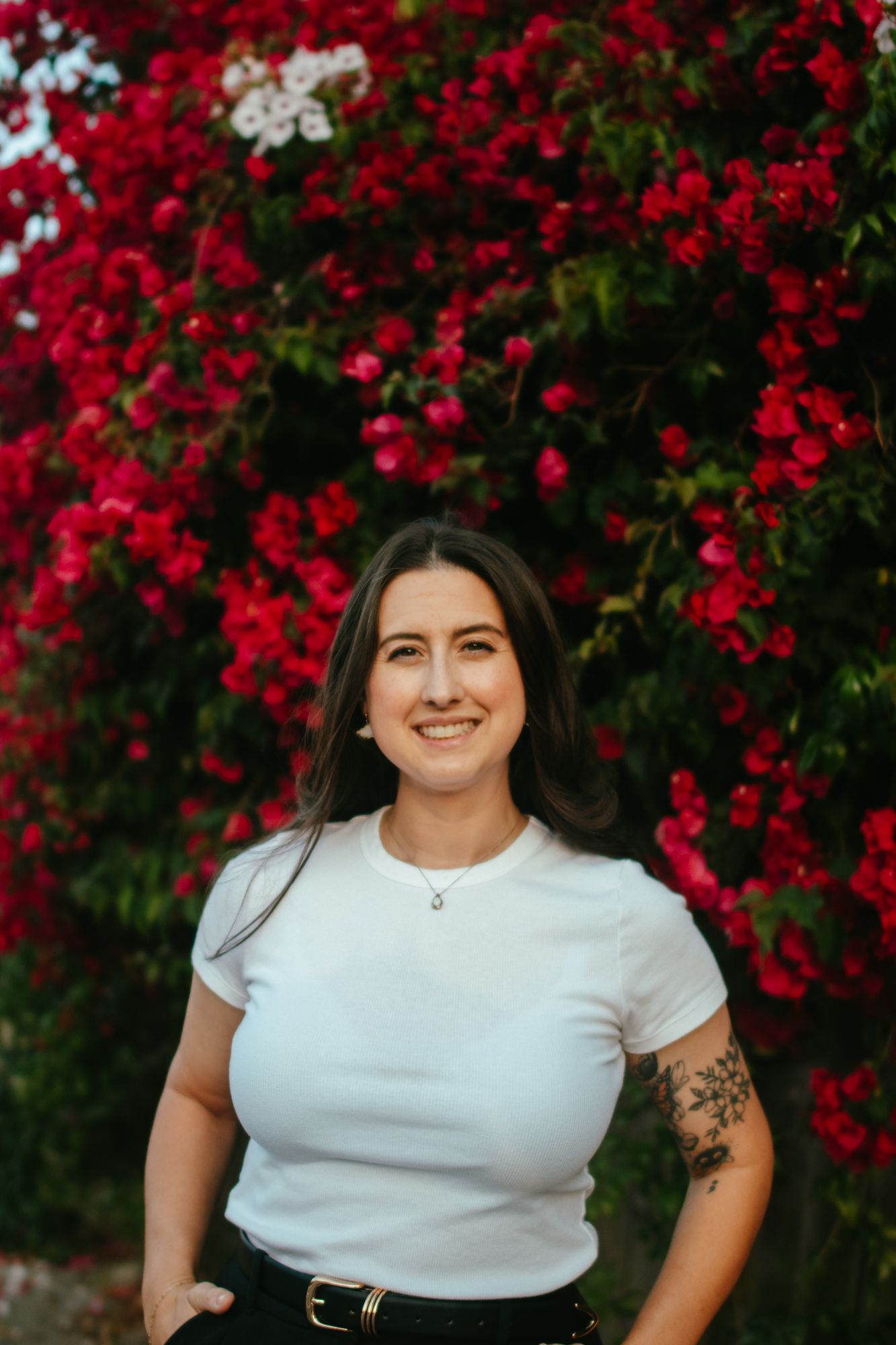 Smiling woman in white t-shirt with arm tattoo standing in front of vibrant red bougainvillea flowers.