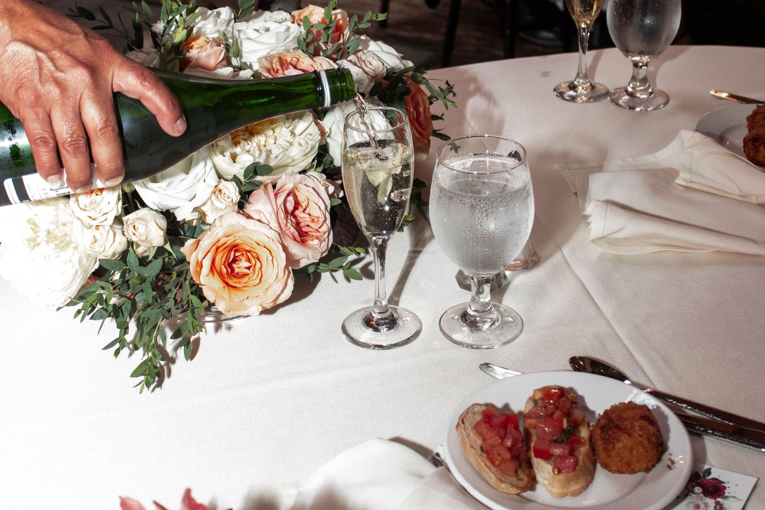 Champagne being poured at wedding reception table with roses, bruschetta appetizers, and crystal glassware.