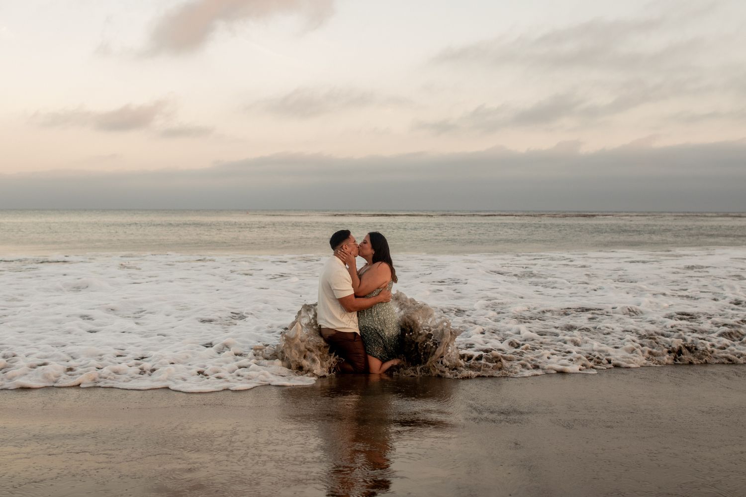 A couple embraces and kisses in the surf on a beach during a golden sunset.