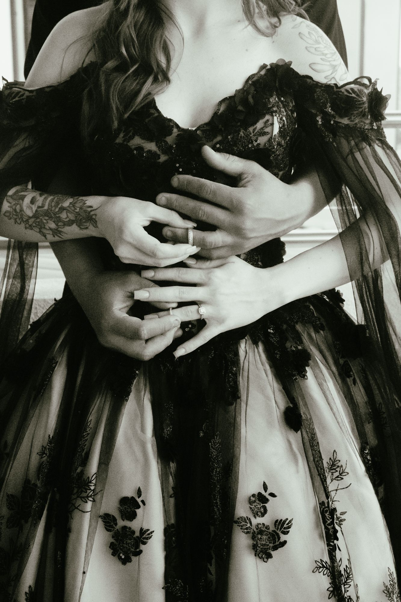 Black and white close-up of couple embracing showing wedding rings in black lace gown.