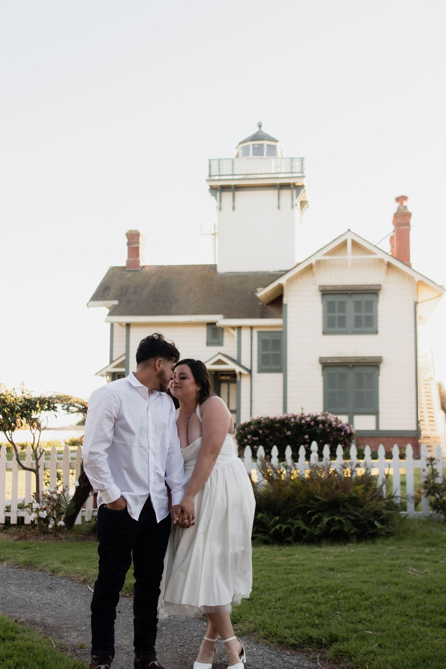 Romantic couple embraces and kisses beside historic white lighthouse with picket fence at dusk.