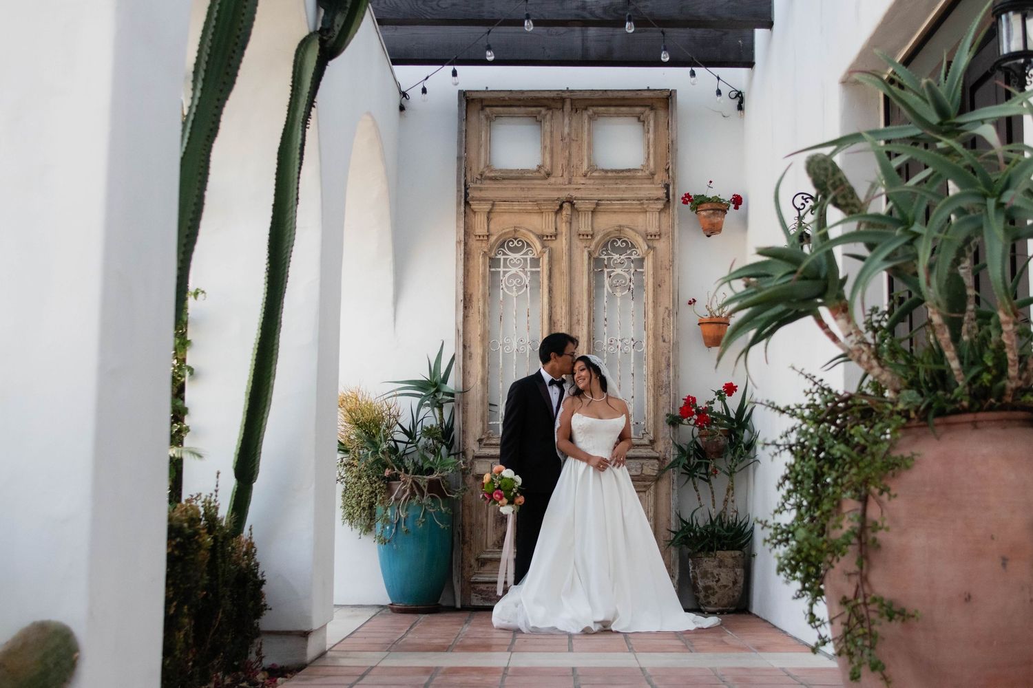 Bride and groom share a romantic moment in front of a rustic wooden door surrounded by lush greenery and cacti.