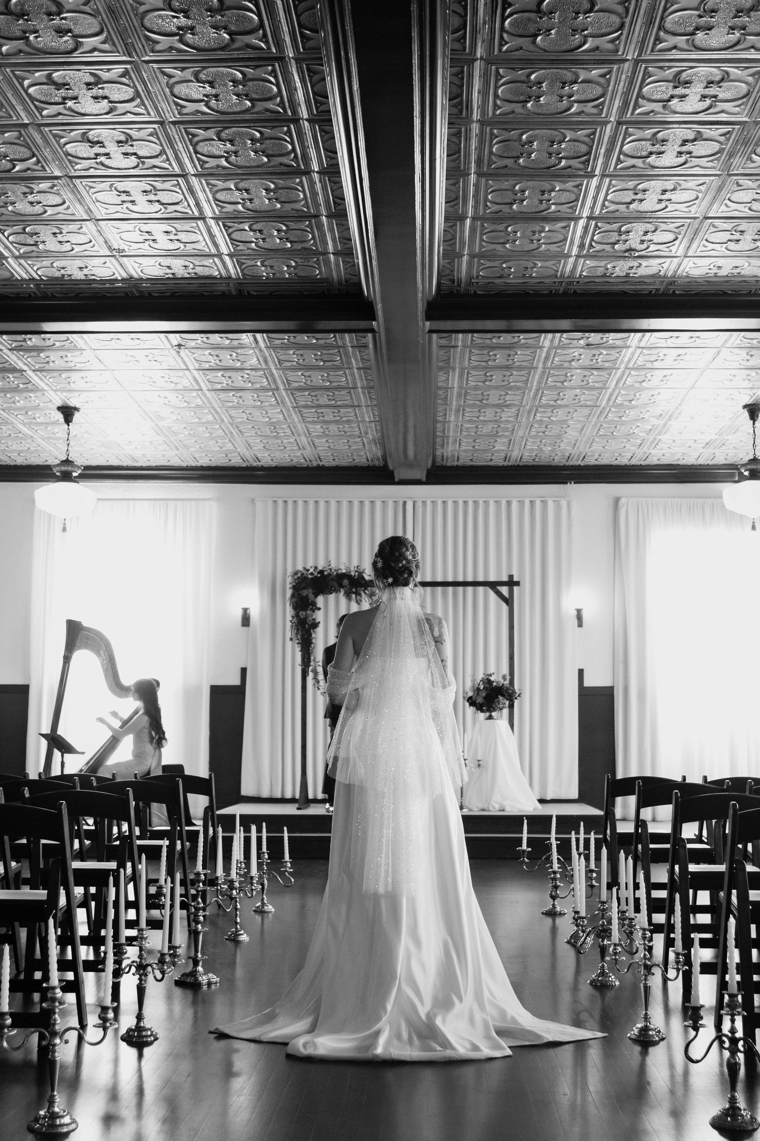 Black and white photo of a bride in a wedding gown standing at the altar of an elegant venue with ornate tin ceilings.