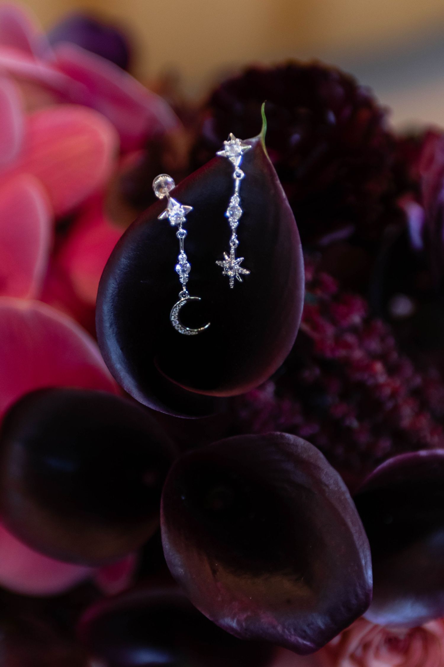 Silver celestial drop earrings with stars and crescent moon displayed on dark burgundy flower petals.