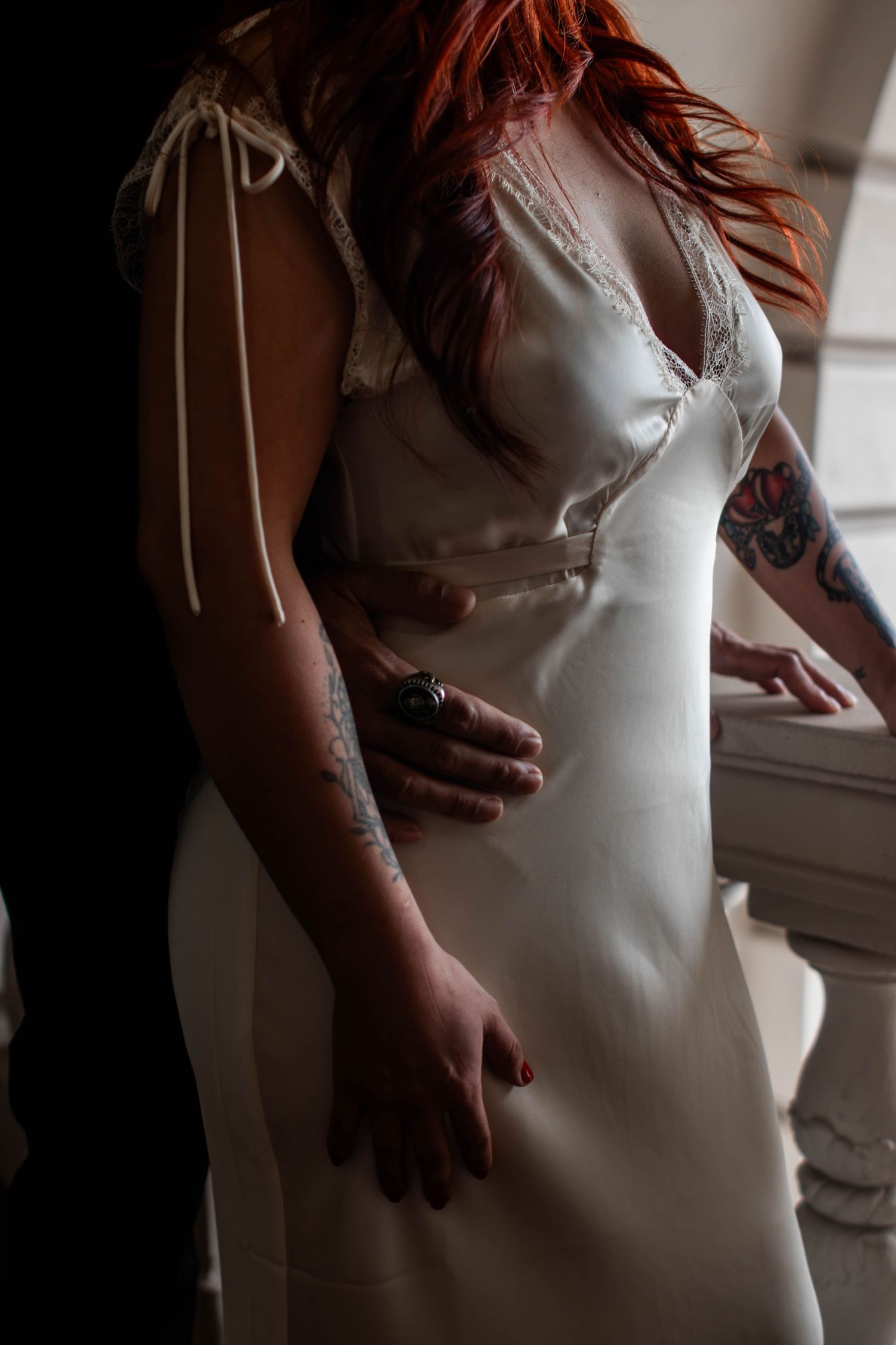 Woman in white satin slip dress with lace trim and colorful arm tattoo poses elegantly indoors.