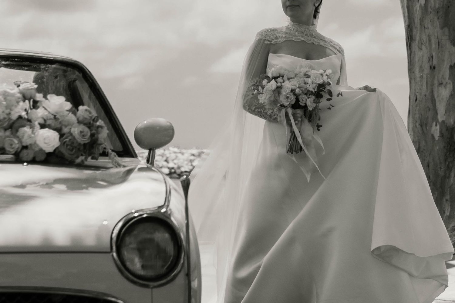 Black and white photo of a bride in a white gown holding a bouquet beside a vintage convertible car.