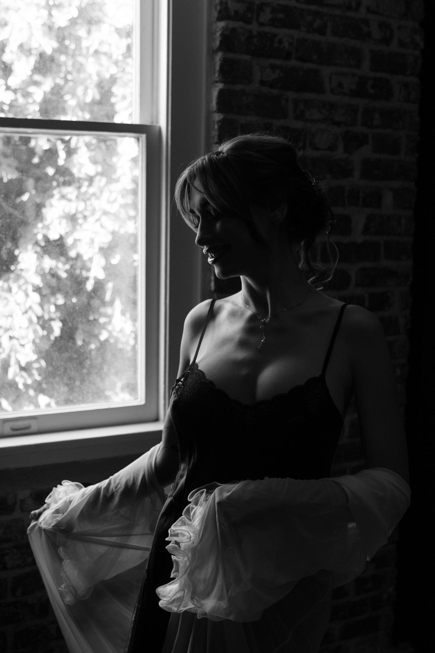Black and white photo of a woman in lingerie and sheer robe standing by a sunlit window near a brick wall.