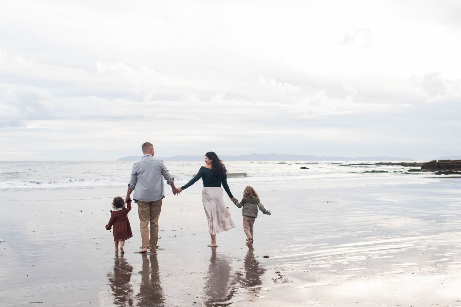 Family of four holding hands walking along a reflective beach shoreline under cloudy skies.
