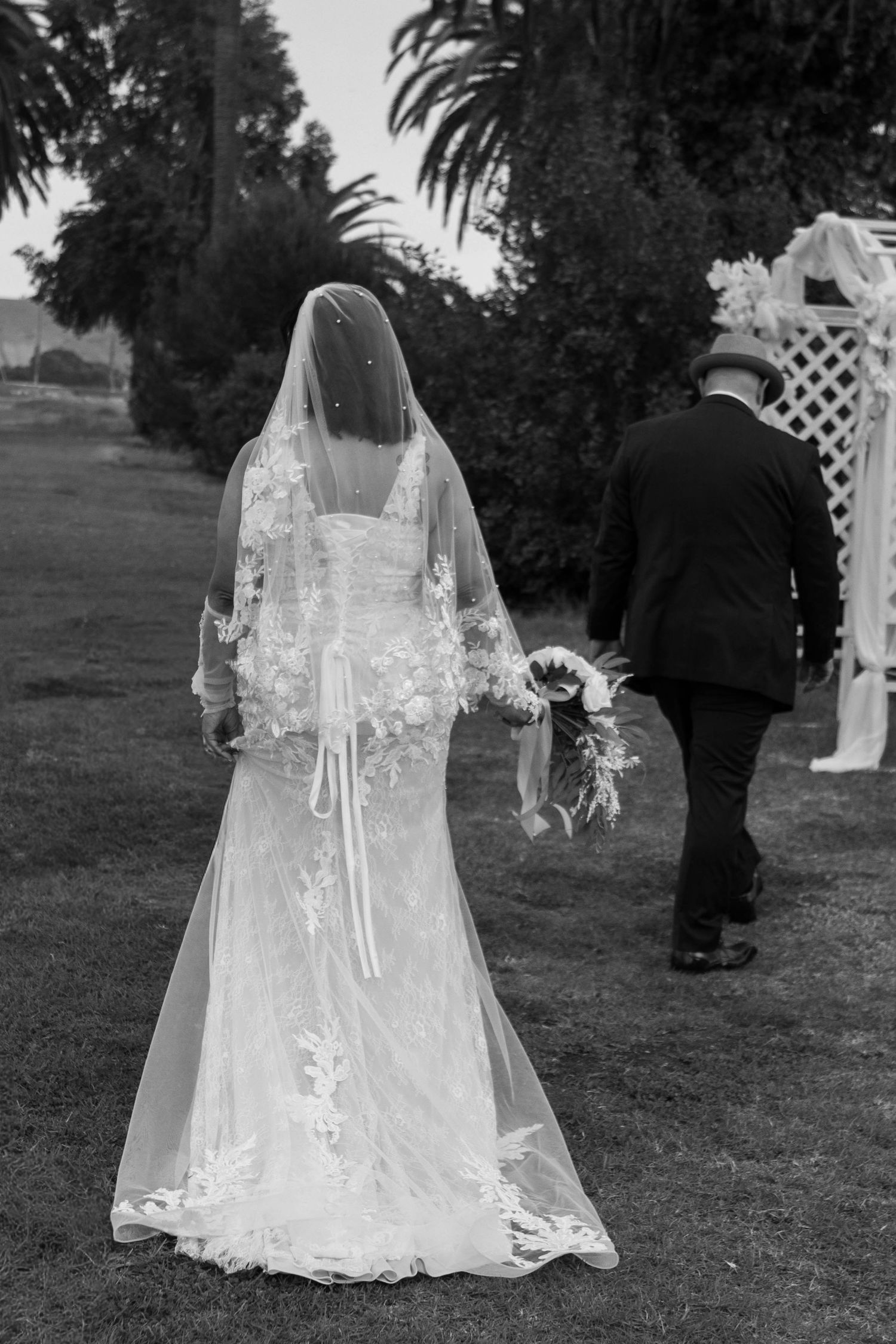 Black and white rear view of bride in lace gown and veil walking with escort toward altar.