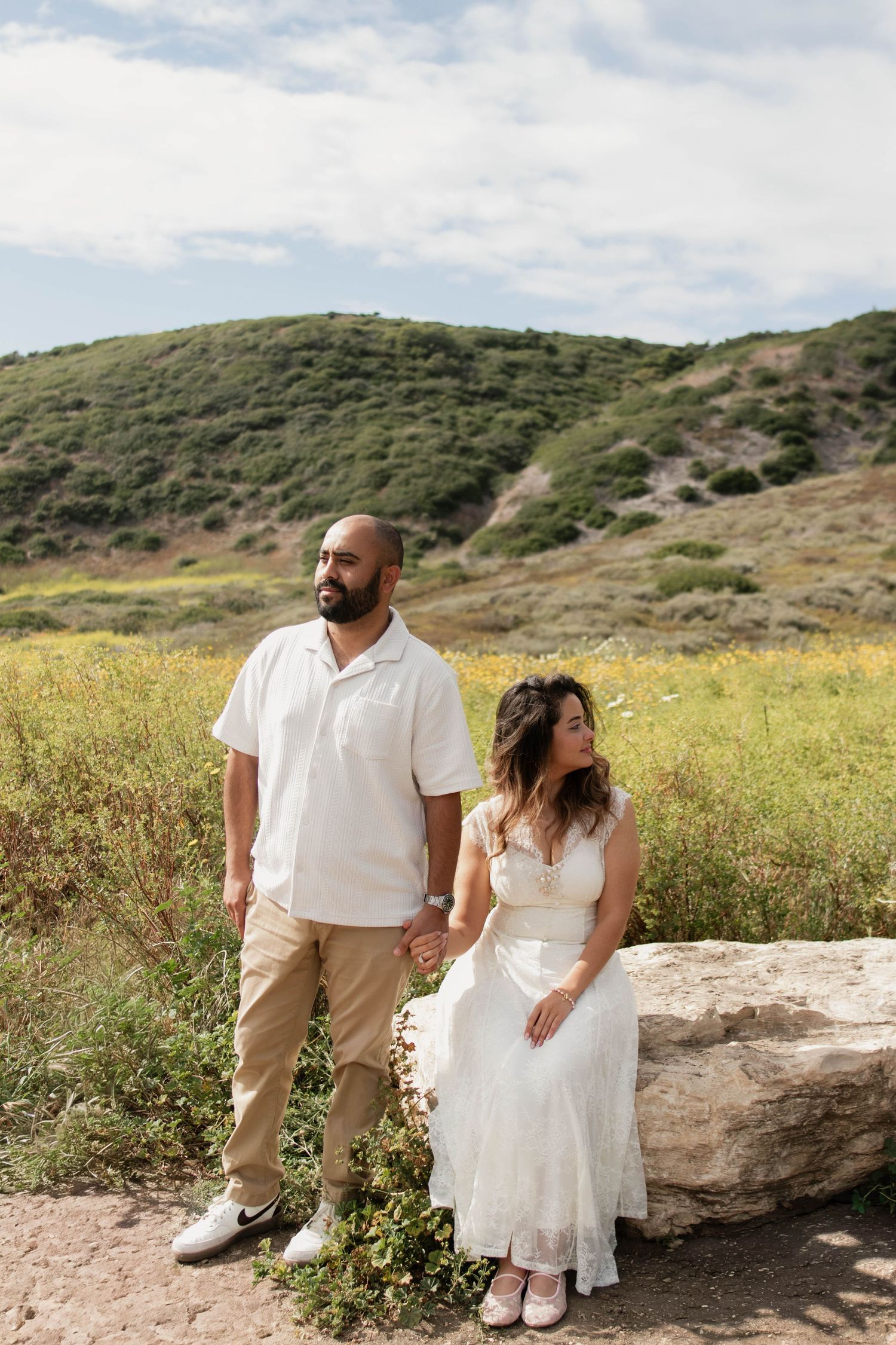 Engaged couple posing on a rock with green hills and wildflowers in the background outdoors.