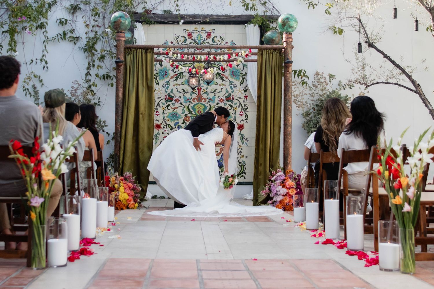 Couple shares first kiss at outdoor wedding ceremony with colorful floral backdrop and rose petal aisle.