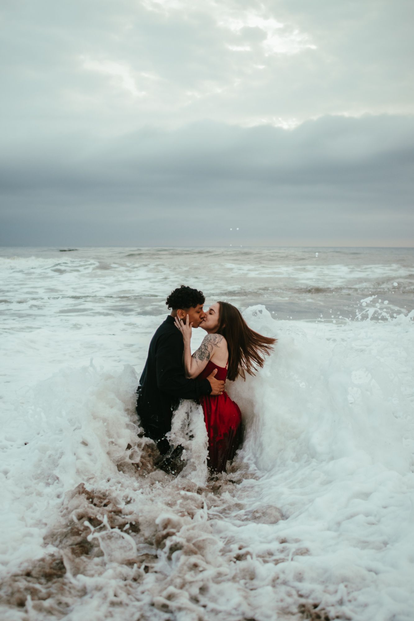 A couple kisses passionately as a large ocean wave crashes around them on the beach.