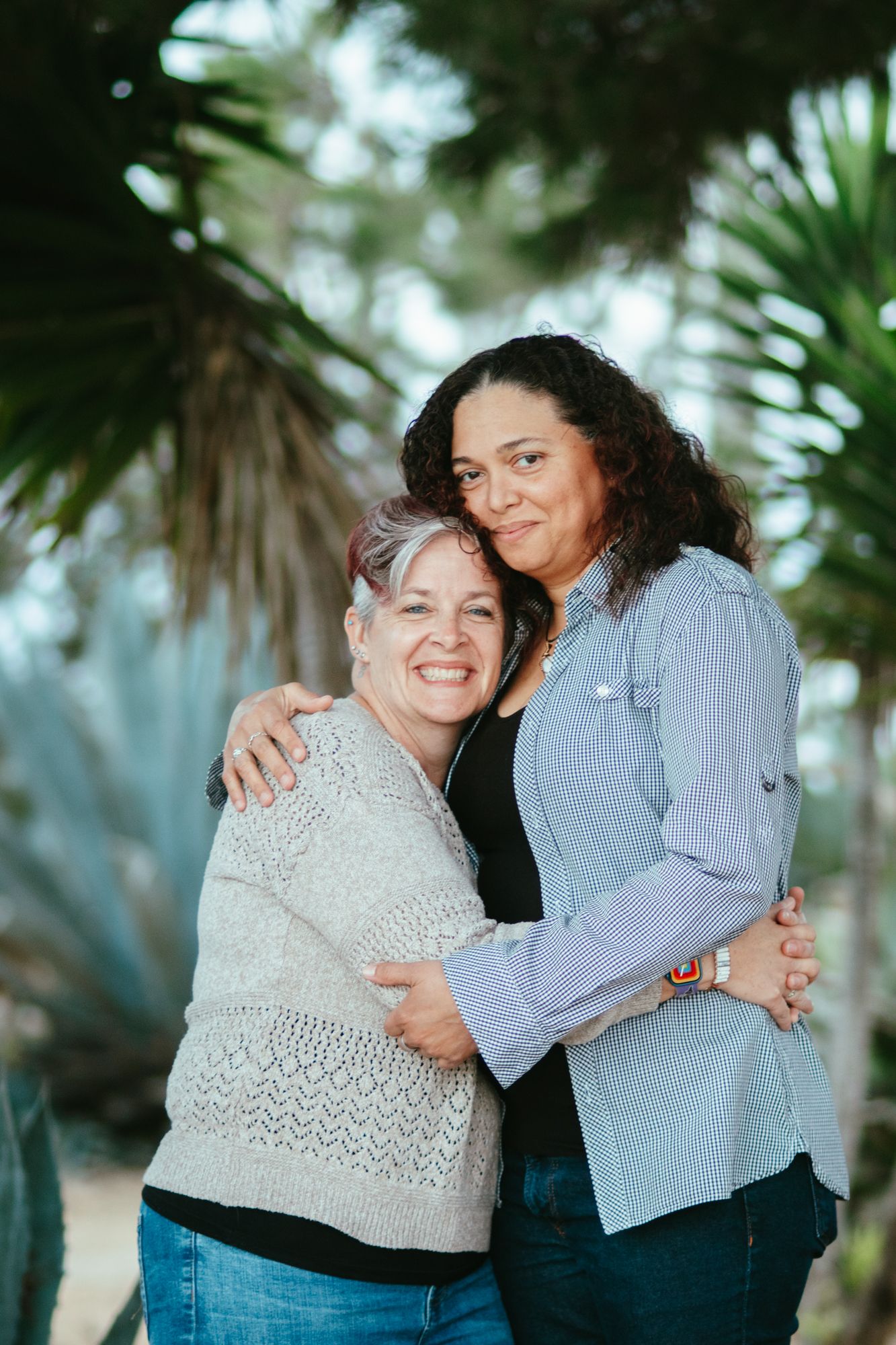 Two women smile and embrace warmly in front of lush tropical palm trees outdoors.