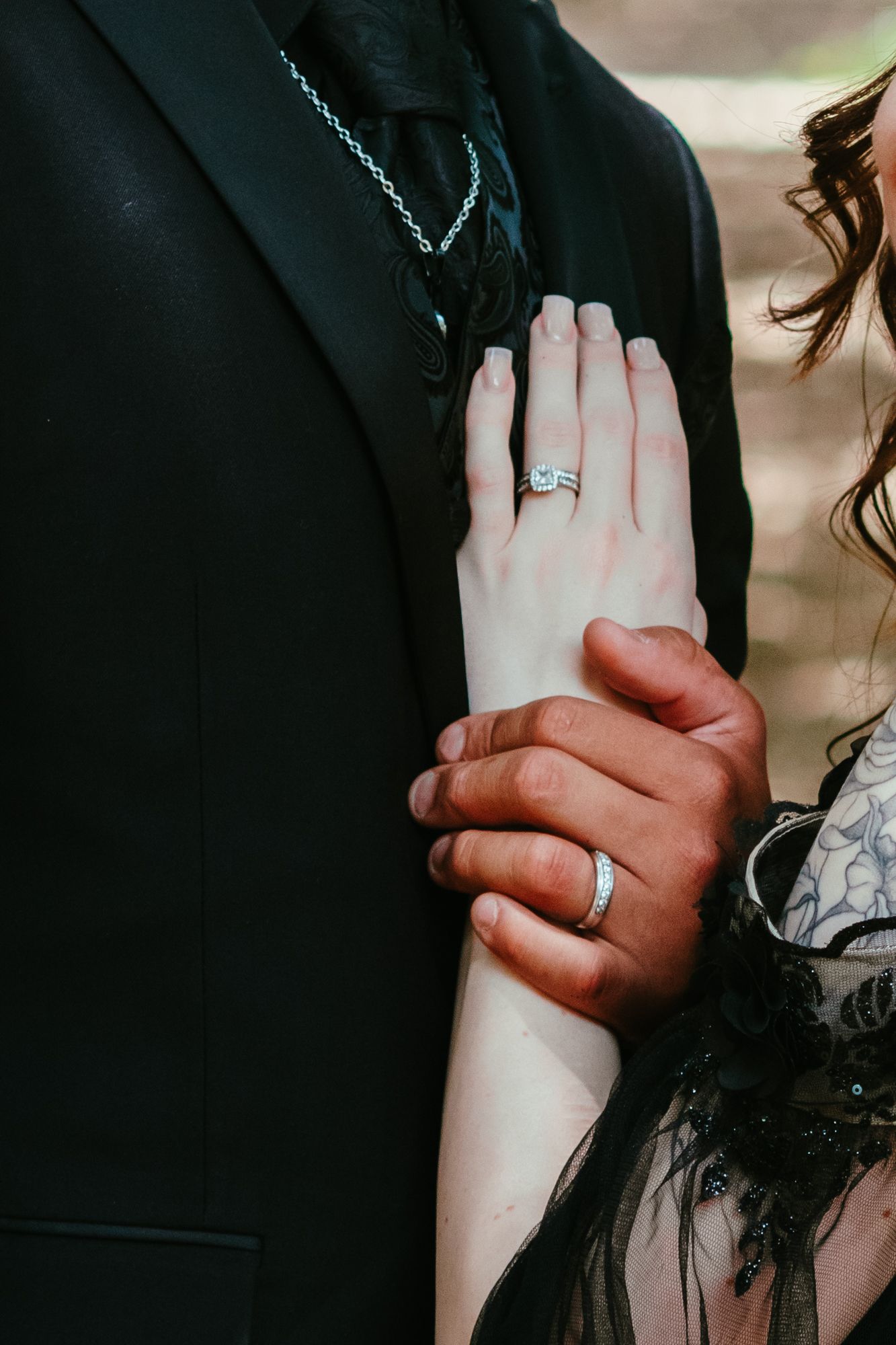 Close-up of interracial couple's hands showing wedding rings, with bride's hand resting on groom's black suit lapel.