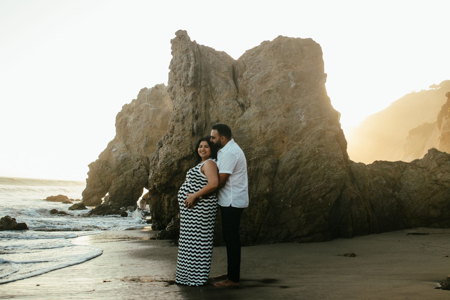 Pregnant couple embracing on beach at sunset with dramatic rock formations behind them.