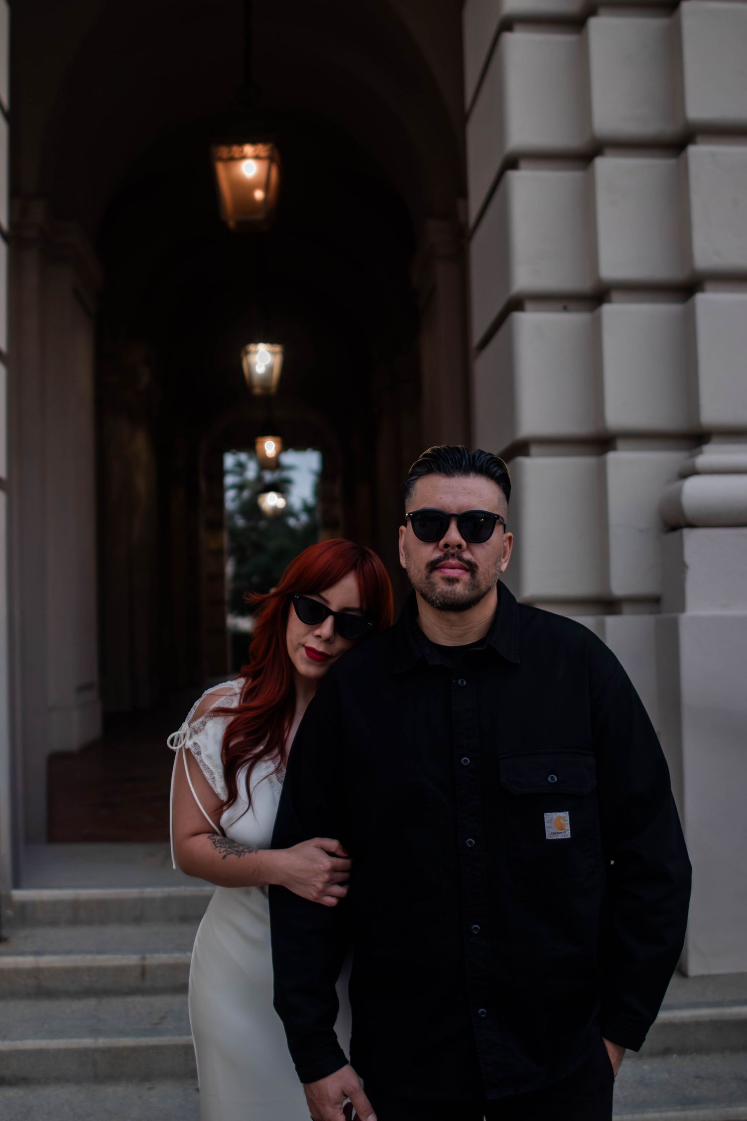 Stylish couple in sunglasses pose outside a classical building with arched columns and warm lighting.